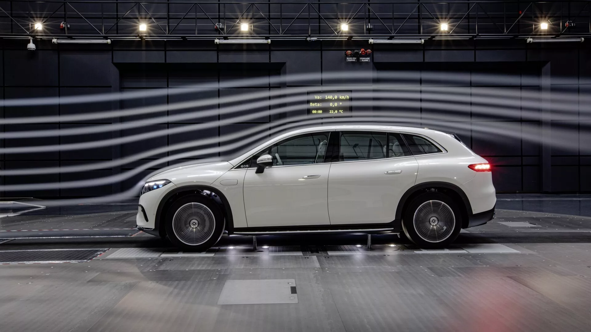 Stranski profil Mercedesa EQS SUV 2022 na izkustveni progi, ki poudarja njegovo elegantno obliko, platišča iz lahke zlitine in odsevni, dobro vzdrževani zunanjost. Sodobni luksuzni crossover SUV ima rahlo nagnjeno streho proti zadnjemu delu, kar nakazuje športno estetiko v kombinaciji s praktičnostjo SUV. Fotografija zajema celotno dolžino avtomobila z zamegljenimi črtami v ozadju, ki simulirajo visoko hitrost, poudarjajoč oblikovanje in domnevne zmogljivosti vozila v nadzorovanem, enobarvnem okolju.