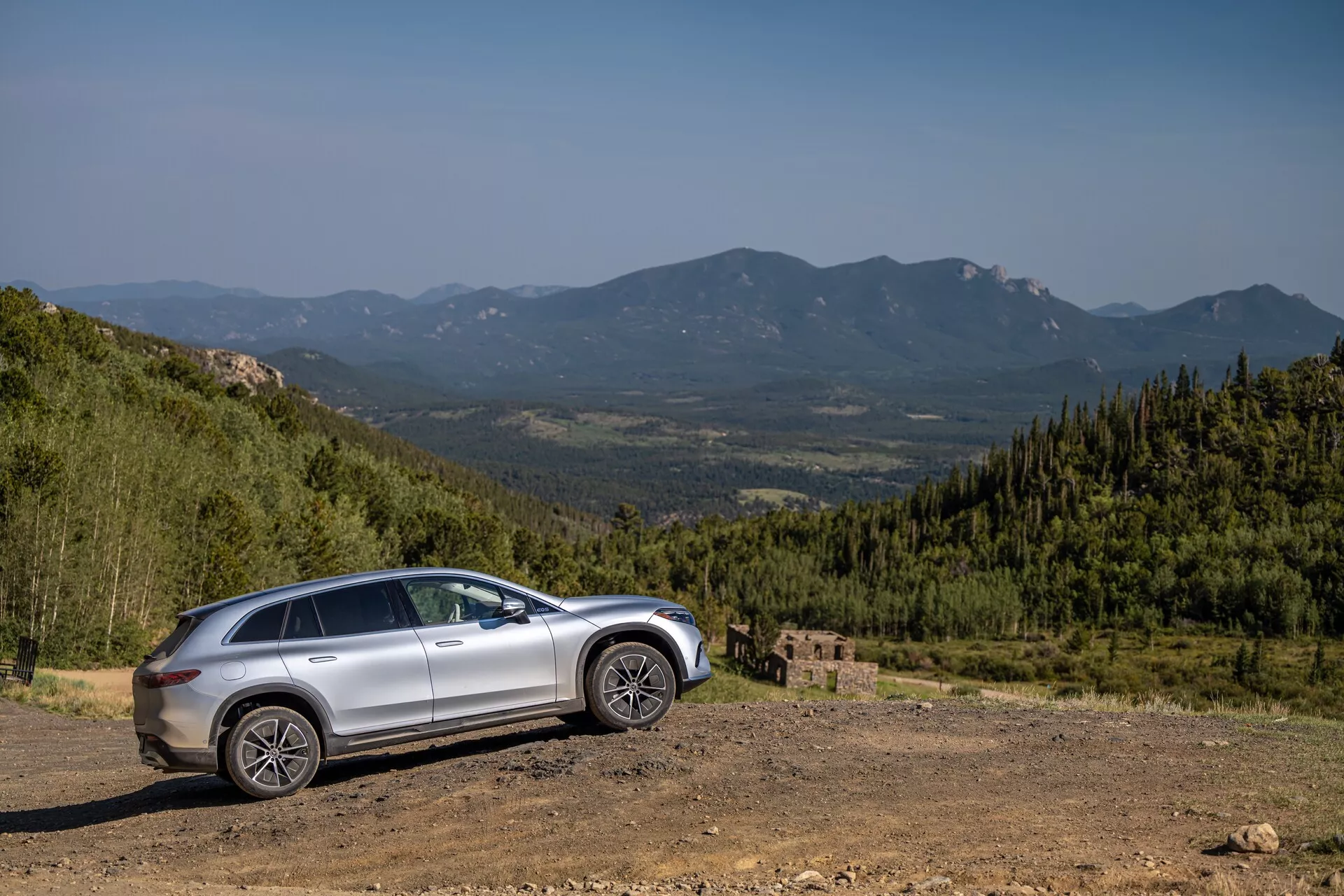 Stranski profil srebrnega Mercedes EQS-SUV iz leta 2022 na neravnem terenu s slikovitim gorskim ozadjem, zajeto pod kotom 30 do 40 stopinj. Značilnosti vključujejo visok odmik od tal, dvignjeno vožnjo, velika robustna kolesa, izklesane linije na vratih, možna strešna okna, bočna ogledala z integriranimi smerniki, zatemnjena okna in sodobne LED žaromete in zadnje luči.