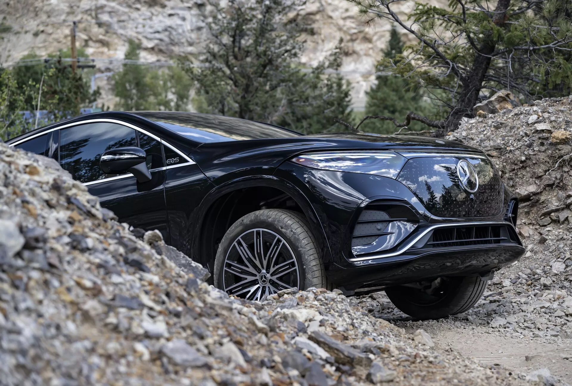 Slika črnega Mercedesa EQS-SUV 2022, ki se premika po terenu s stranskim nizkim profilom. Osredotočen je na prednjo četrtino vozila, prikazuje podrobnosti, kot so sijajna črna površina, elegantne linije, sprednja maska z emblemom, značilni žarometi, mišičasti blatniki, aluminijasta platišča in tekalna plast pnevmatik, primerna za vse terene. Sprednje kolo je rahlo zavrnjeno v desno, kar kaže na manever. Naravno ozadje s kamni in redko vegetacijo poudarja terenske zmogljivosti SUV-ja.