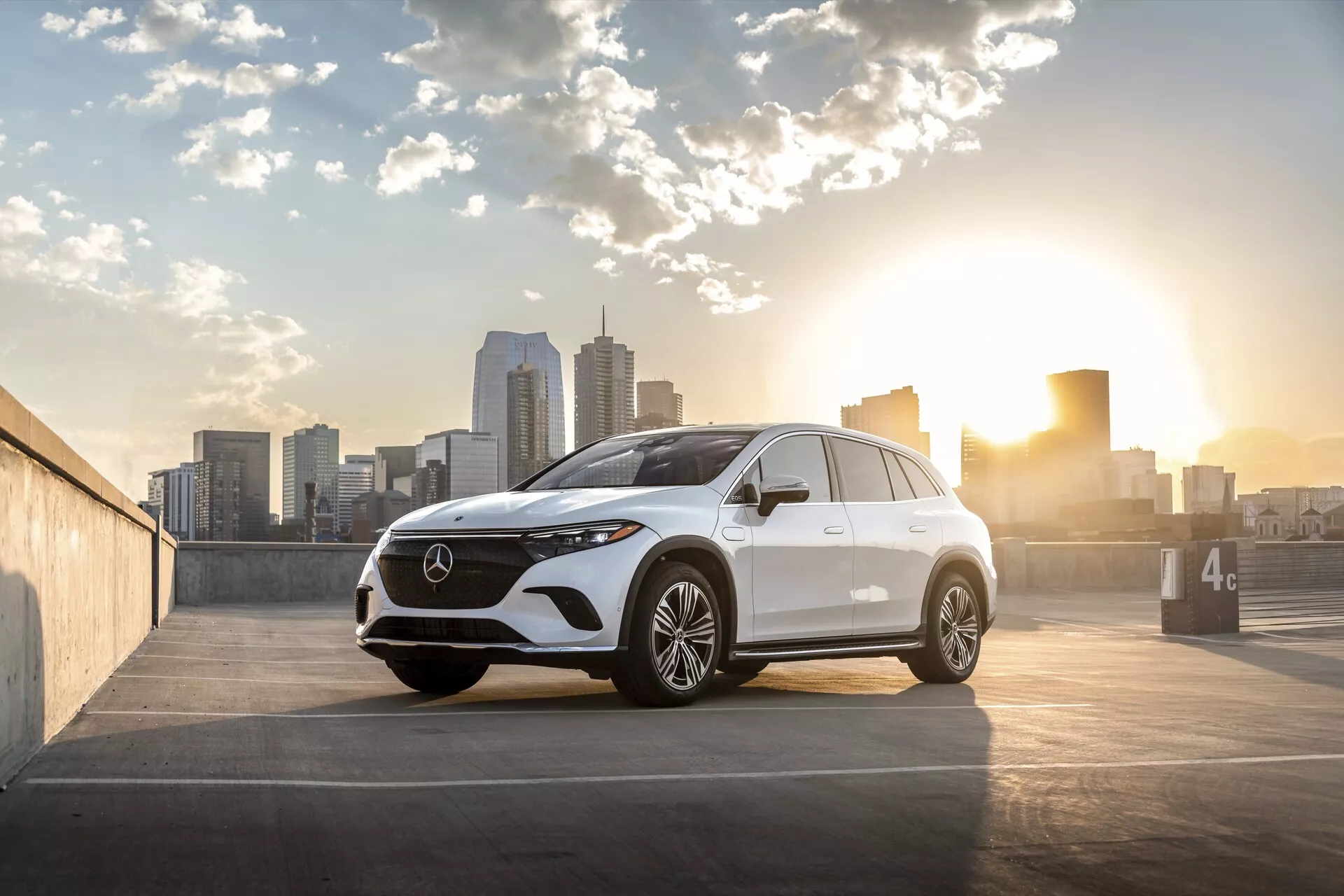 Slika prikazuje sodoben Mercedes EQS-SUV 2022, ki je parkiran na odprtem parkirišču z mestnim obzorjem v ozadju, ujet ob sončnem vzhodu ali zahodu s toplo osvetlitvijo. Pogled s treh četrtin spredaj poudarja mrežo, žaromete, zasnovo sprednjega odbijača, pokrov motorja in voznikovo stran, posnet iz nizkega kota blizu nivoja koles. Podrobnosti vključujejo vidno mrežo z velikim emblemom, elegantne LED žaromete, sprednjo kamero ali skupino senzorjev, večkraka aluminijasta platišča, vgrajene smerne kazalnike na stranskih ogledalih, nežne obrisne linije za aerodinamiko in rahlo padajočo linijo strehe. Sestava fotografije postavi avto na desno stran okvira, z mestnim obzorjem in soncem na levi, dodajajoč bleščanje in artistično privlačnost, primerno za avtomobilske, luksuzne in mestno življenjske kontekste.