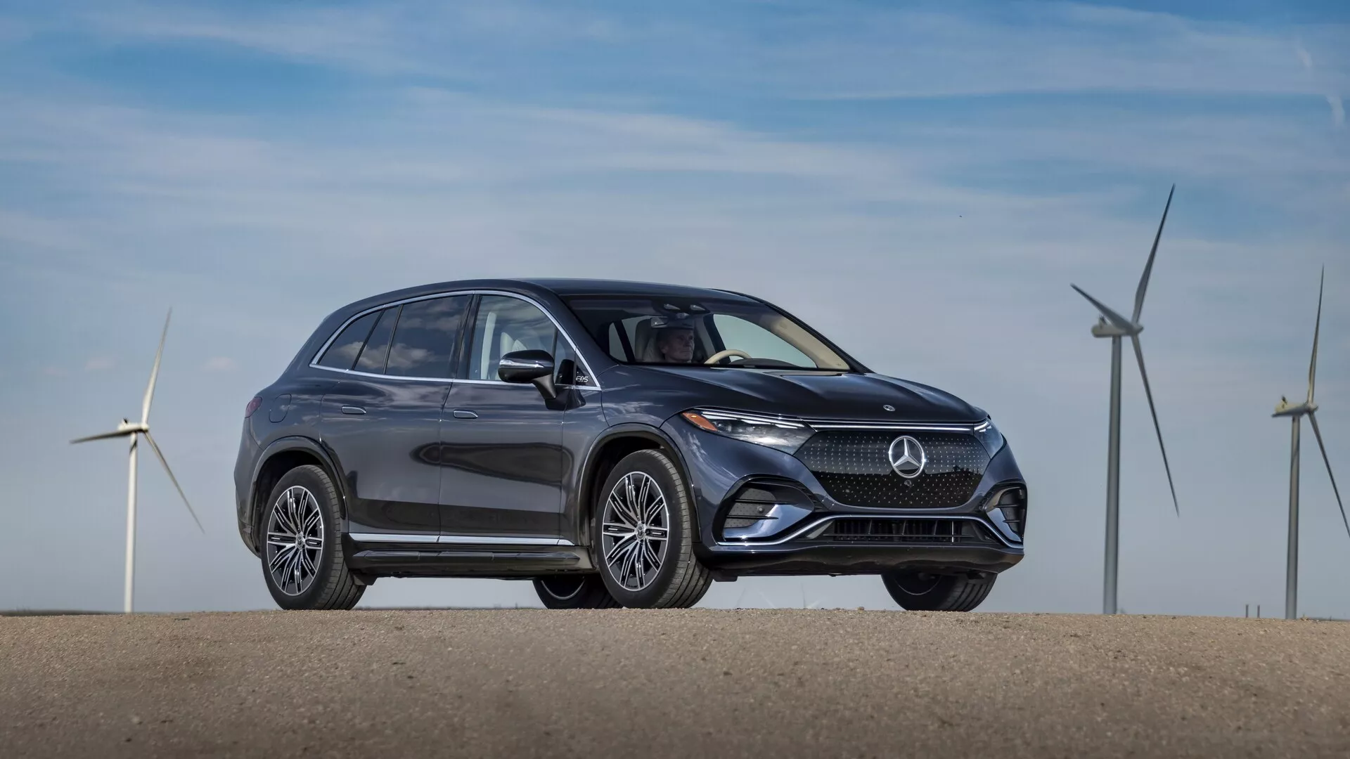 Stranski profil temno obarvanega SUV Mercedes EQS 2022, parkiranega na enakomerni površini. Slika, posneta z rahlim kotom sprednje četrti, prikazuje eleganten in sodoben dizajn z zaobljenimi linijami in gladkimi površinami, ki poudarjajo aerodinamično učinkovitost. Opazne funkcije vključujejo izrazito rešetko z osrednjim logotipom blagovne znamke, elegantne LED žaromete, zračne vhode ali senzorje v spodnjem odbijaču, prefinjena  večkraka lita platišča, stranska ogledala z integriranimi smerniki, kromirano obrobo oken in rahlo padajočo strešno linijo z nosilci na strehi. V ozadju so vetrne turbine, kar namiguje na ekološko ali električno naravo vozila.