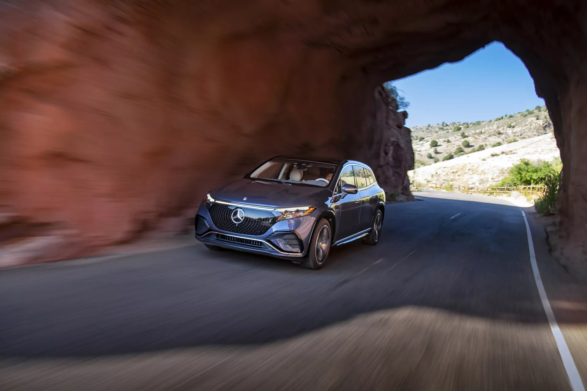 Slika avta Mercedes EQS-SUV 2022, ki se pojavi iz tunela na cesti, posneta iz nizkega zornega kota, s poudarkom na sprednji levi strani, prikazuje moderni dizajn vozila s izrazito rešetko, logotipom, LED dnevnimi lučmi, večkrakimi aluminijastimi platišči in kovinsko barvo. Avto je v gibanju, kar kažejo zamegljeno ozadje in kolesa. Okolica prikazuje skalnat teren z redko vegetacijo, kar nakazuje gorsko ali puščavsko območje v jasnem vremenu.