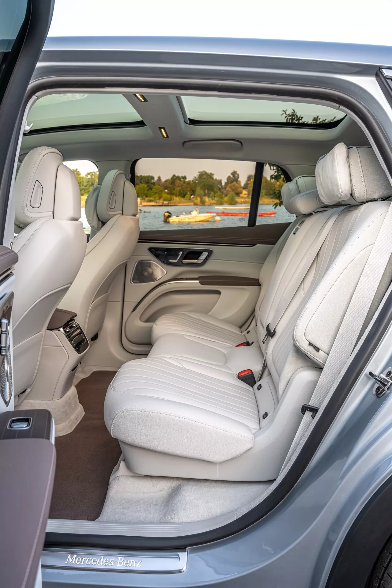 Pogled v notranjost Mercedes EQS SUV 2022 iz zadnjih desnih vrat, ki prikazuje razkošno zadnjo klop iz lahke kremne barvne usnje s tremi vzglavniki, panoramsko streho, elektronske kontrole, individualne klimatske zračne šobe, kromirane poudarke, mrežo zvočnikov, žep za shranjevanje in kakovostno preprogo.