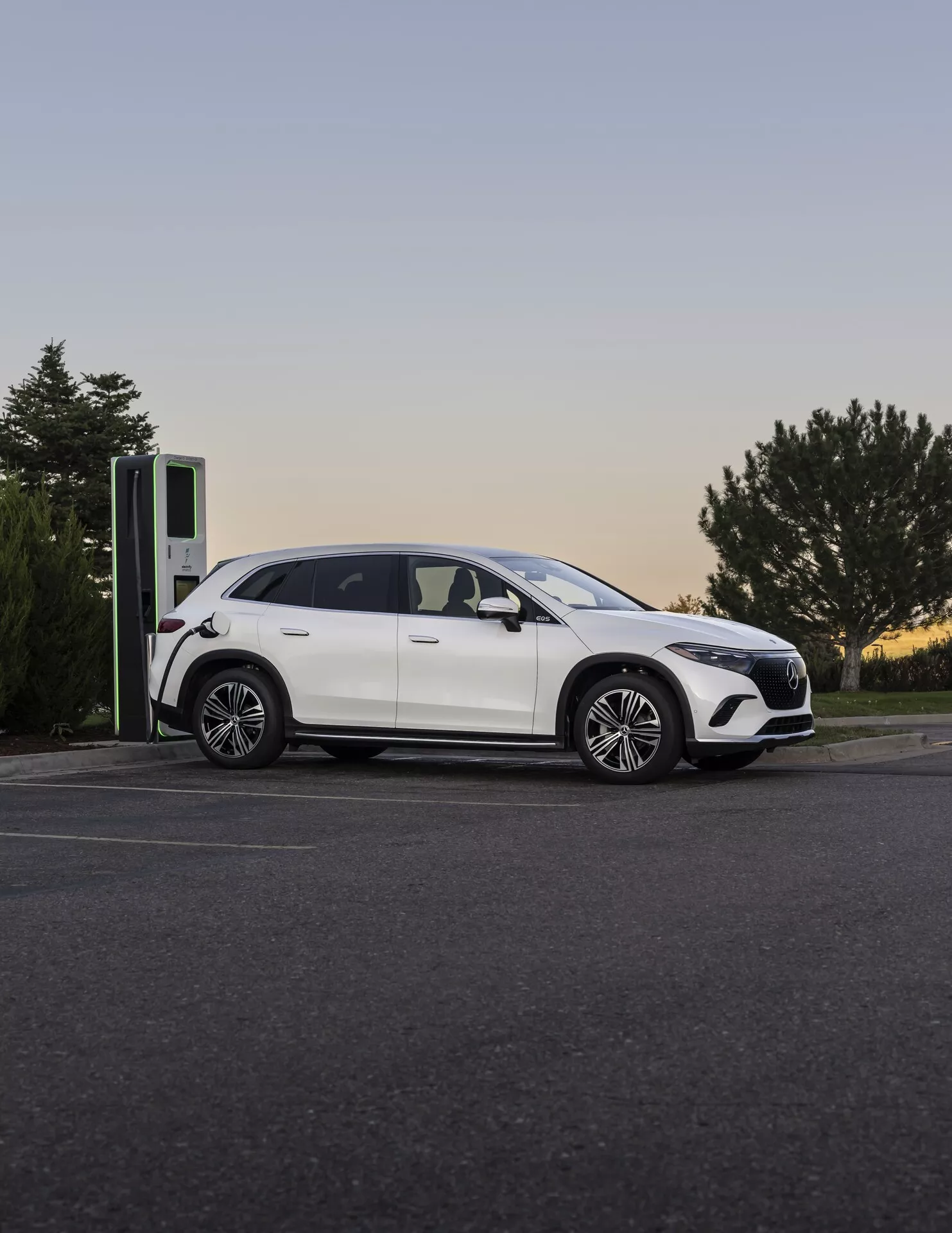 Sodoben Mercedes EQS-SUV 2022 pri polnilni postaji v mraku, fotografiran iz stranskega sprednjega kota, ki poudarja njegov eleganten profil, lita platišča in polnilni priključek.