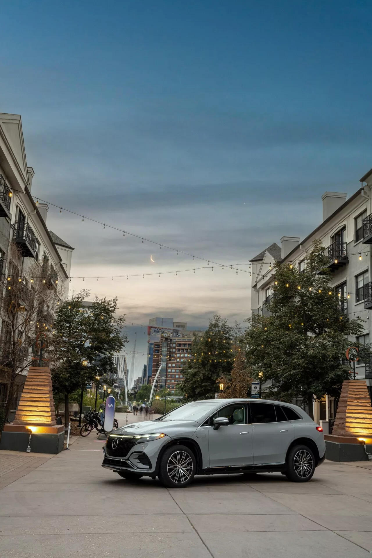 Sprednji trikotni pogled na srebrni Mercedes EQS-SUV 2022, ki poudarja njegovo glavno rešetko z emblemom, dnevne LED luči, lita platišča in karoserijo. Avtomobil je parkiran v luksuznem mestnem okolju z dekorativno razsvetljavo in zgradbami, kar poudarja njegov luksuzni in sodobni dizajn.