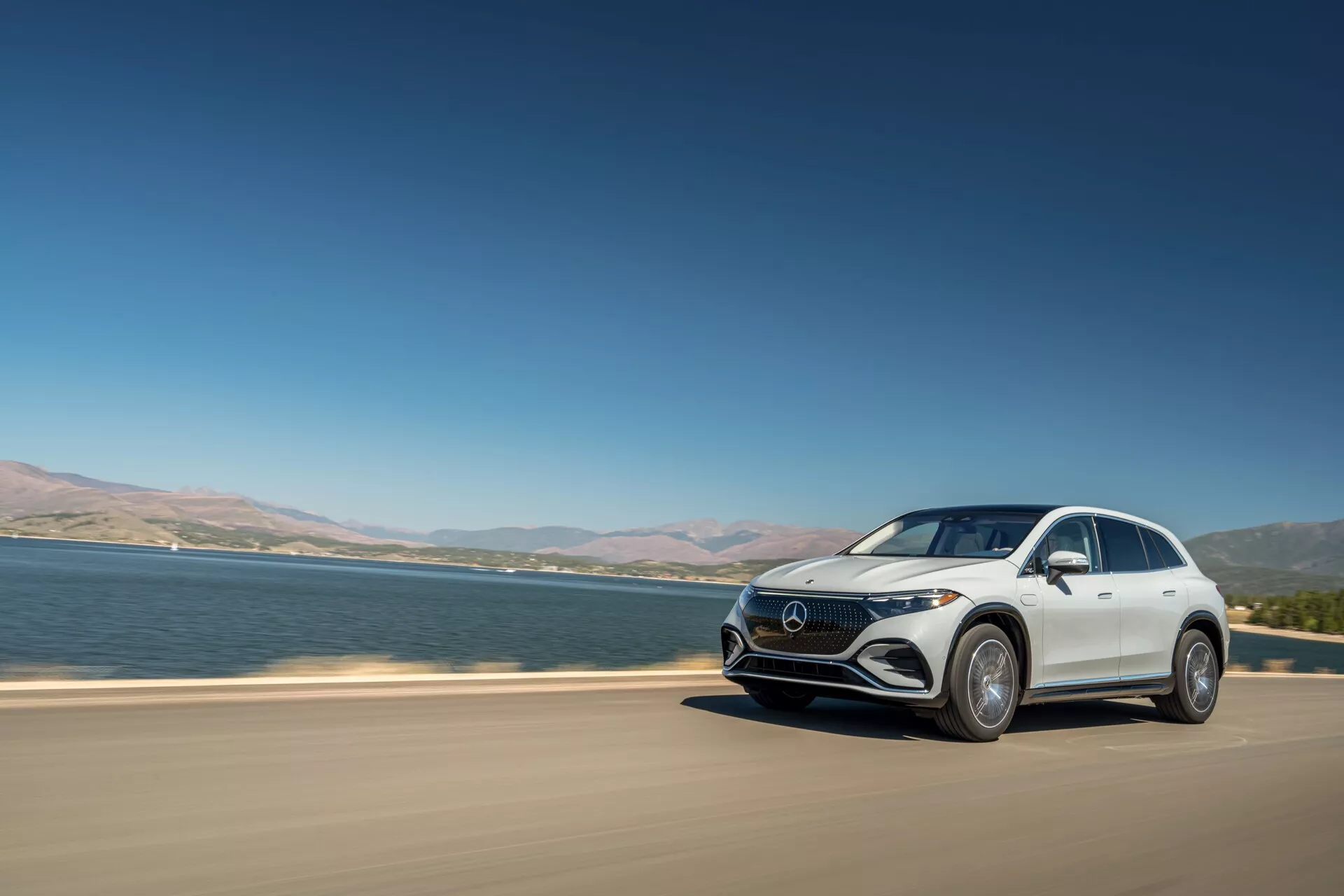 Moderen luksuzni crossover SUV, Mercedes-Benz EQS, ki vozi po asfaltirani cesti v bližini velikega vodnega telesa. Slika prikazuje avto iz treh četrtin sprednjega pogleda v močni dnevni svetlobi, poudarjajoč značilnosti, kot so LED žarometi, značilna rešetka, aerodinamična oblika in velika lita platišča. Vozilo je v gibanju, kar nakazuje dinamiko, z jezerom ali reko v ozadju in čistim nebom, kar je značilno za premium električni model.