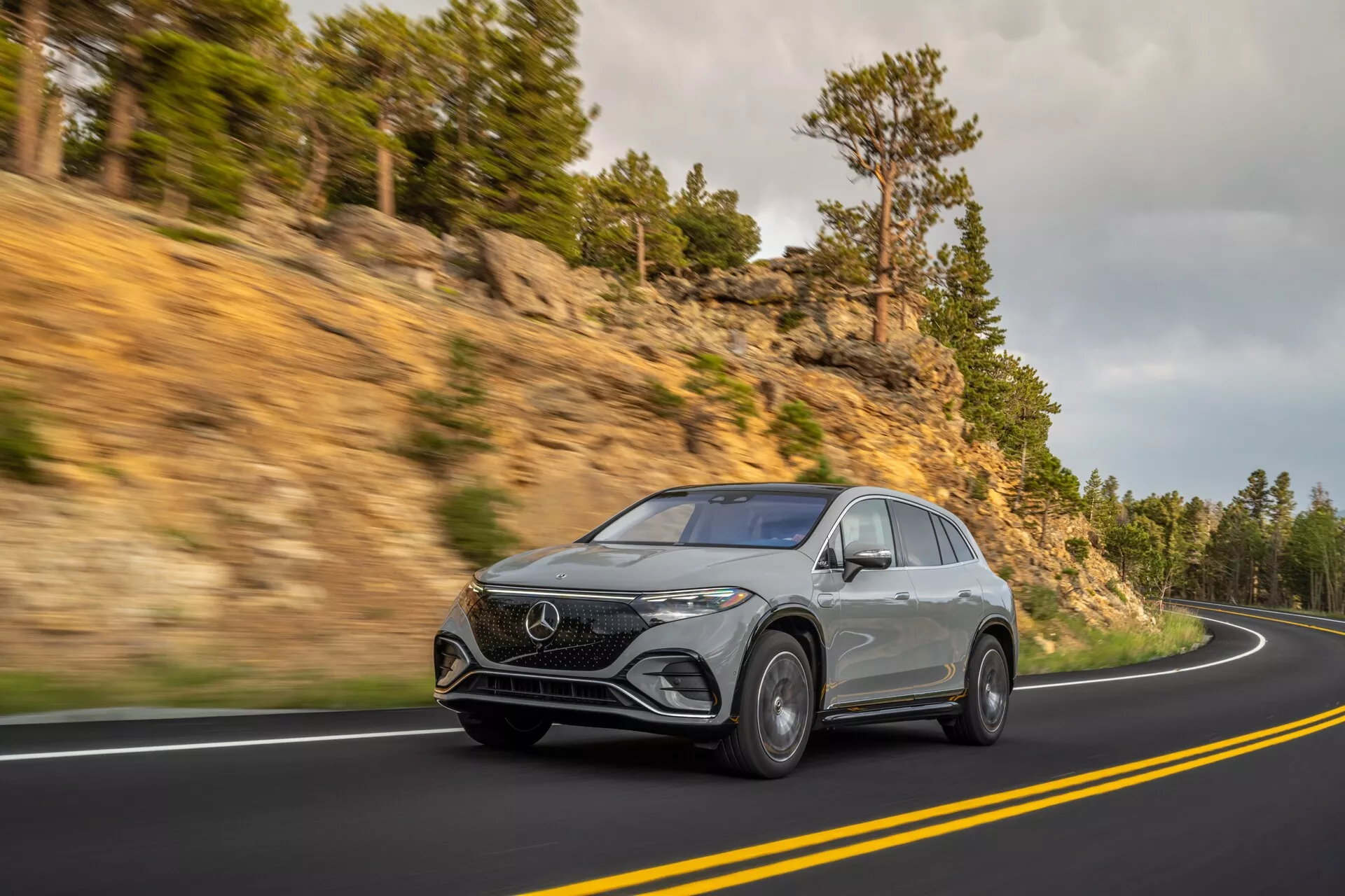 Dinamičen posnetek srebrnega Mercedes EQS SUV 2022 na ovinkasti gorski cesti, posnet iz treh četrtin spredaj, da poudari njegovo višino in prisotnost na cesti. S sodobnimi LED žarometi, značilnim sprednjim znakom, luksuznimi aluminijastimi platišči in aerodinamičnimi linijami, ki kažejo na sodoben dizajn.