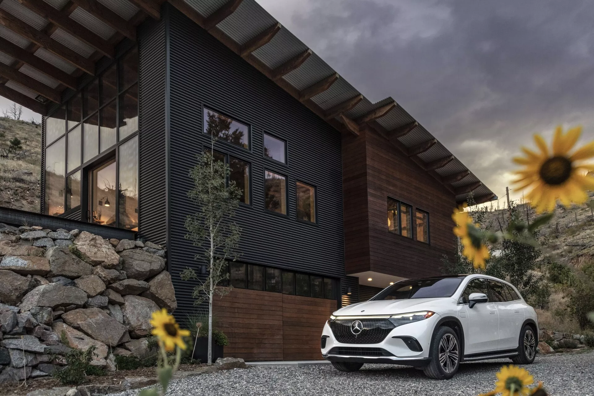 Slika luksuznega SUV-ja Mercedes-Benz EQS 2022, parkiranega pred sodobno hišo z oblačnim nebom v ozadju. Posnetek iz nizkega kota poudarja držo in velikost vozila, osredotoča se na sprednjo rešetko, sklop žarometov, sprednji odbijač, konture pokrova motorja in voznikovo stran, ki se razteza do zadnjih vrat. Značilnosti vključujejo LED dnevne luči in elegantna kolesa, ki prispevajo k občutku razkošja in napredne tehnologije. Beli SUV kontrastira s temno hišo in skalnato pokrajino, obdan je z rahlo zamegljenimi cvetovi zaradi majhne globinske ostrine, kar poudarja SUV kot glavno osrednjo točko.