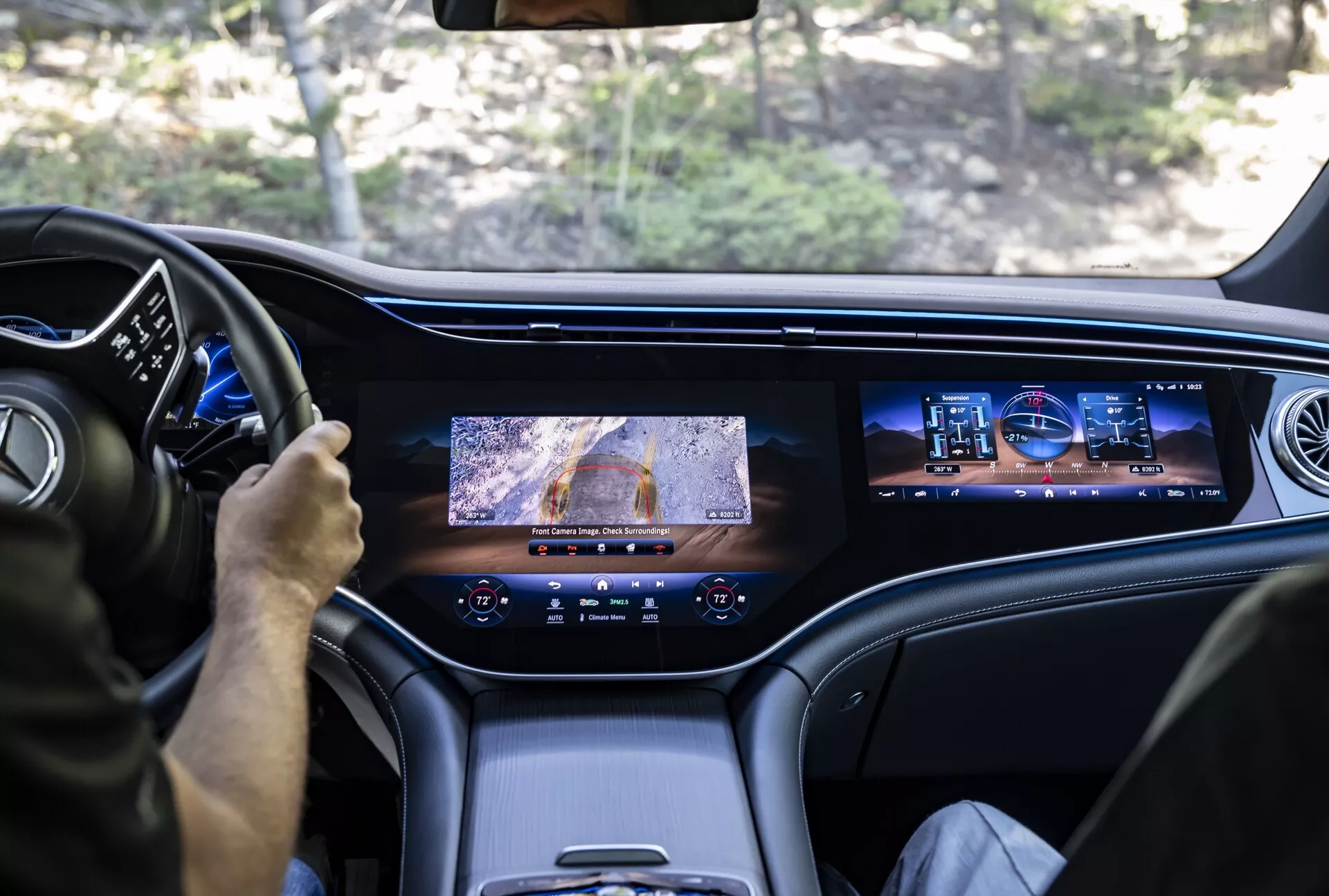 Pogled z zadnjega sedeža Mercedes EQS-SUV 2022, ki prikazuje notranji prednji del; prikazuje voznika, ki uporablja večfunkcijski volan, digitalno armaturno ploščo, velik osrednji zaslon z pogledom kamere, sekundarni zaslon za infotainment za potnika, vrhunske usnjene ali visoko kakovostne materiale kabine in moderno oblikovana zračnika, poudarjajoč luksuzne in napredne tehnološke lastnosti vozila.