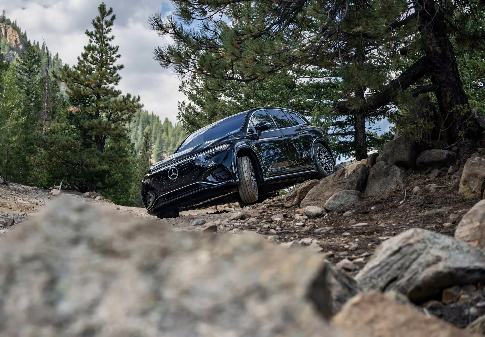 Posnetek črnega SUV-ja Mercedes-Benz EQS 2022 iz nizkega kota, proti surovemu zunanjemu ozadju, ki prikazuje njegovo sprednjo levo stran s parcialnim pogledom na desno strani. Slika izpostavlja značilne žaromete, masko z Mercedesovim znakom, aluminijasta platišča in robustne pnevmatike na makadamski poti s skalami, poudarjajoč terensko zmogljivost in luksuzni dizajn.