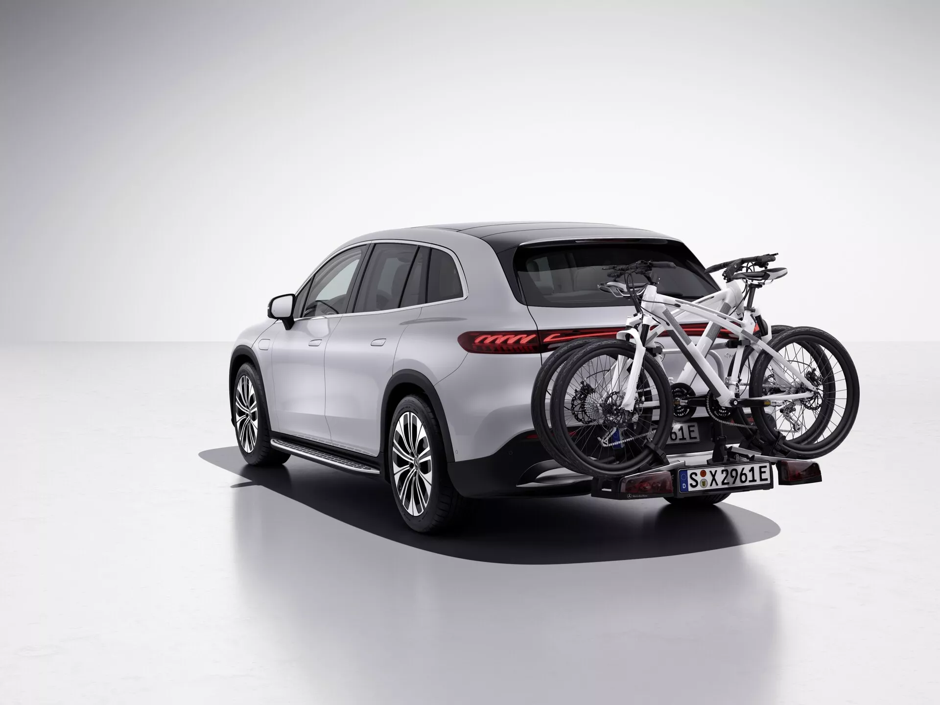 Slika prikazuje SUV Mercedes EQS 2022 v studijskem okolju s čisto, belo ozadje, gledano z zadnje tričetrtinske perspektive, ki prikazuje sopotnikovo stran. Vozilo je opremljeno z nosilcem za kolesa na zadnjem vlečnem priključku, ki drži eno kolo. SUV ima eleganten, sodoben dizajn z gladkimi linijami, značilen zadnji svetlobni sklop z vodoravnimi rdečimi LED zadnjimi lučmi, večkrakimi aluminijastimi platišči in svetlo barvo karoserije, verjetno srebrna ali siva, v kontrastu s črnim spodnjim trimom in oblogami kolesnih obročev. Stranski ogledali, strešni nosilci in obrobe oken so v temnejšem tonu s panoramsko streho, ki dodaja prestižen občutek. Ta slika poudarja funkcije kot so LED osvetlitev, športni dizajn koles in nosilec za kolesa, kar jo naredi primerno za kategorije kot so Vozila, Avtomobilska tehnologija, Športna oprema/zunanji pripomočki in Luksuzni SUV-ji.