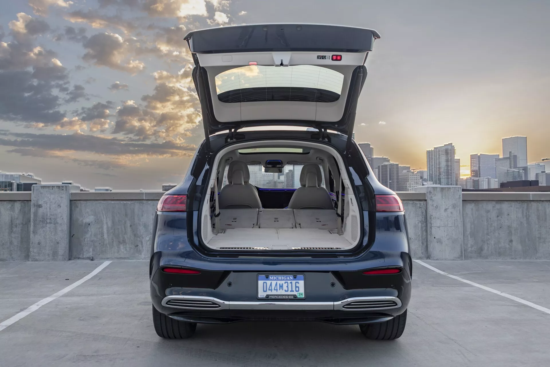 Pogled od zadaj na Mercedes EQS-SUV 2022, posnet pod rahlo nizkim kotom, s poudarkom na odprtem prtljažniku. Avto je parkiran na zgornji etaži parkirne strukture, z odprtim nebom in oddaljenimi stavbami. Fotografija zajema celotno širino zadnjega dela, vključno z zadnjimi lučmi, zadnjim odbijačem in delom zadnjih koles. Okoliška osvetlitev in pastelno obarvano nebo nakazujeta, da je bila slika posneta v zgodnjih večernih urah ali ob mraku. Slika poudarja tovorni prostor vozila, zasnovo zadnjih luči, reflektorje in odpiranje prtljažnika, pri čemer se opira na naravno osvetlitev za tople tone in mehke sence.