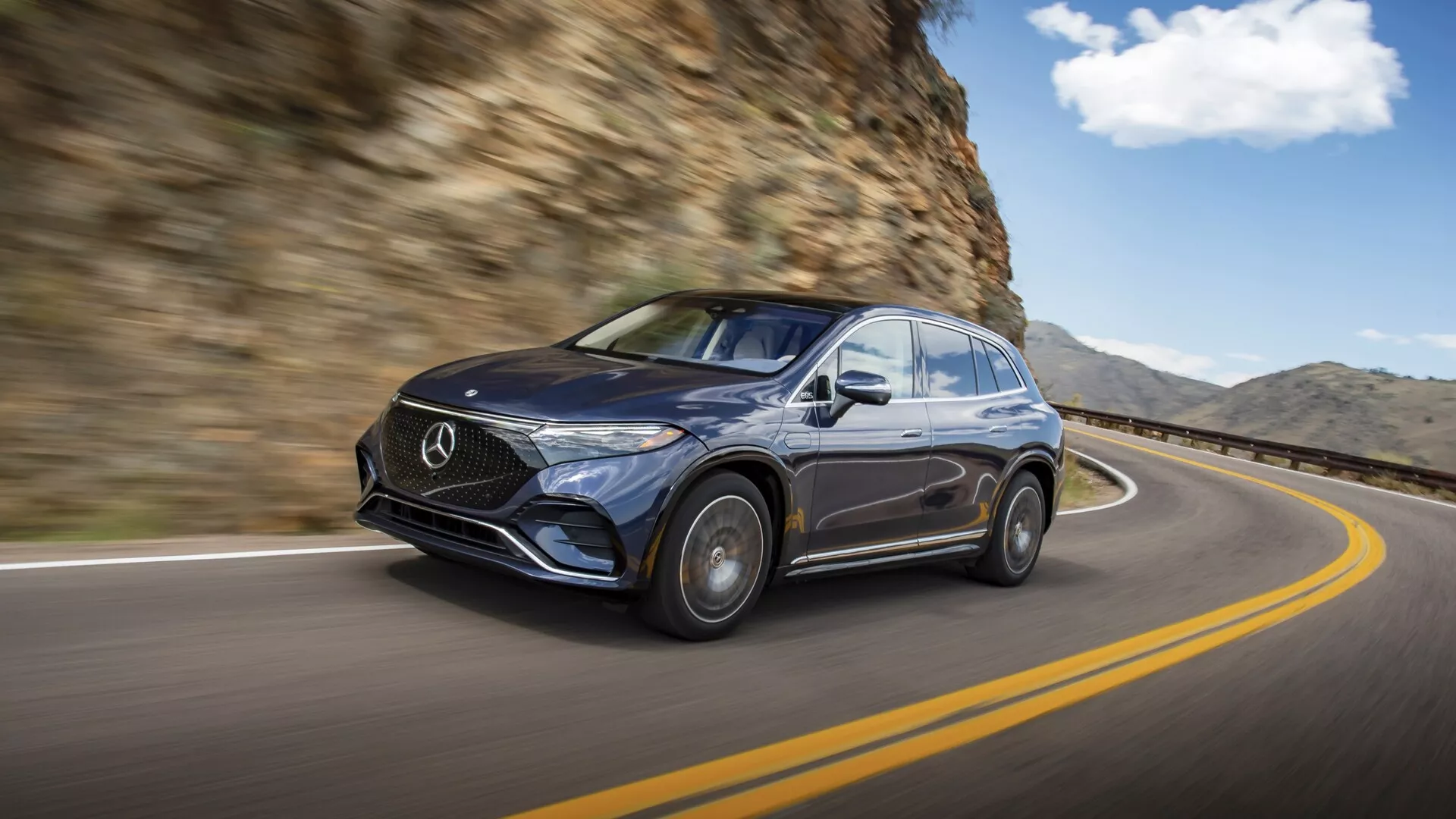 Slika temnega Mercedes EQS SUV 2022 v gibanju, posneta iz sprednjega levega tričetrtinskega pogleda. Avto se vozi po zaviti cesti s skalno steno na desni strani. Poudarek je na luksuzni zasnovi z izrazito mrežo z Mercedesovim emblemom, elegantnimi žarometi, sijočo površino in deloma vidnimi aluminijastimi platišči. Zamegljeno ozadje poudarja občutek gibanja, poudarjajoč vozne sposobnosti in vrhunsko estetiko vozila v naravnem voznem okolju.