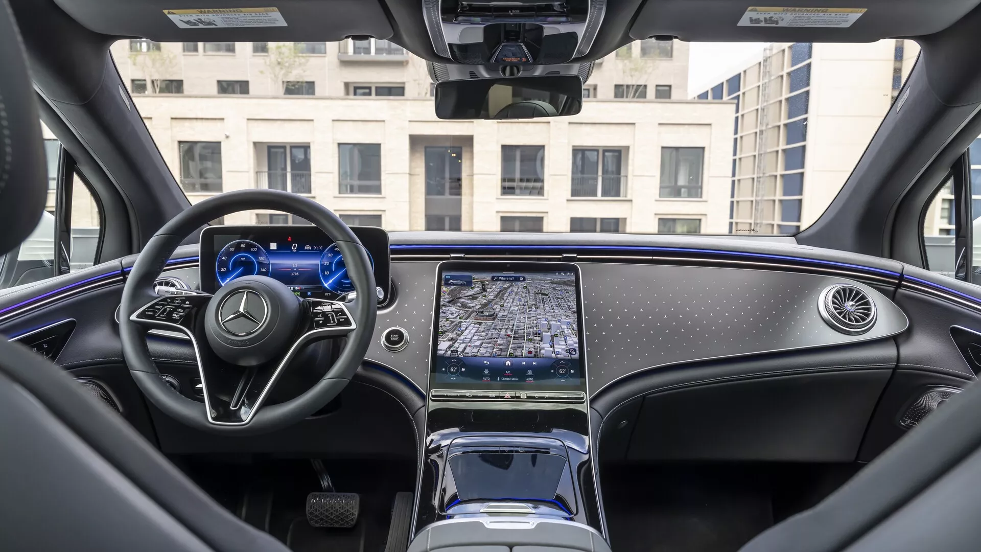 Pogled v notranjost Mercedes EQS SUV 2022 iz perspektive zadnjega sedeža, ki prikazuje armaturno ploščo, večnamenski volan z ravnim spodnjim delom in kovinskimi poudarki ter osrednjo konzolo z podolgovatim zaslonom na dotik infozabavnega sistema. Digitalna instrumentna plošča je osvetljena, minimalistični elektronski menjalnik, shranjevalni predali in držala za pijačo pa so vidni. Razkošna notranjost avtomobila vključuje visokokakovostne materiale, polirane lesene obloge in delno vidno panoramsko streho, kar ponazarja sodoben avtomobilski dizajn in napredne tehnološke funkcije.