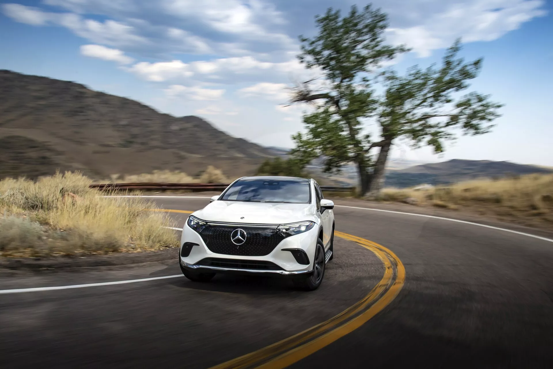 Dinamičen pogled s sprednje tričetrtine na Mercedes EQS-SUV 2022 v gibanju, ki prikazuje oblikovanje rešetke, žarometov, pokrova motorja, kolesa in stranskih kontur. Avto zavzema desno stran okvirja in se premika v levo, z zamegljenim ozadjem, ki nakazuje gibanje. Vidne funkcije vključujejo LED dnevne luči, obliko žarometov, odbijač z vgrajenimi zračnimi odprtinami ali senzorji ter aerodinamično oblikovana stranska ogledala. Cesta in naravna krajina nakazujeta uporabo na avtocesti ali za potovanja, kar poudarja zmogljivost, razkošje in potencialno napredno tehnologijo vozila.