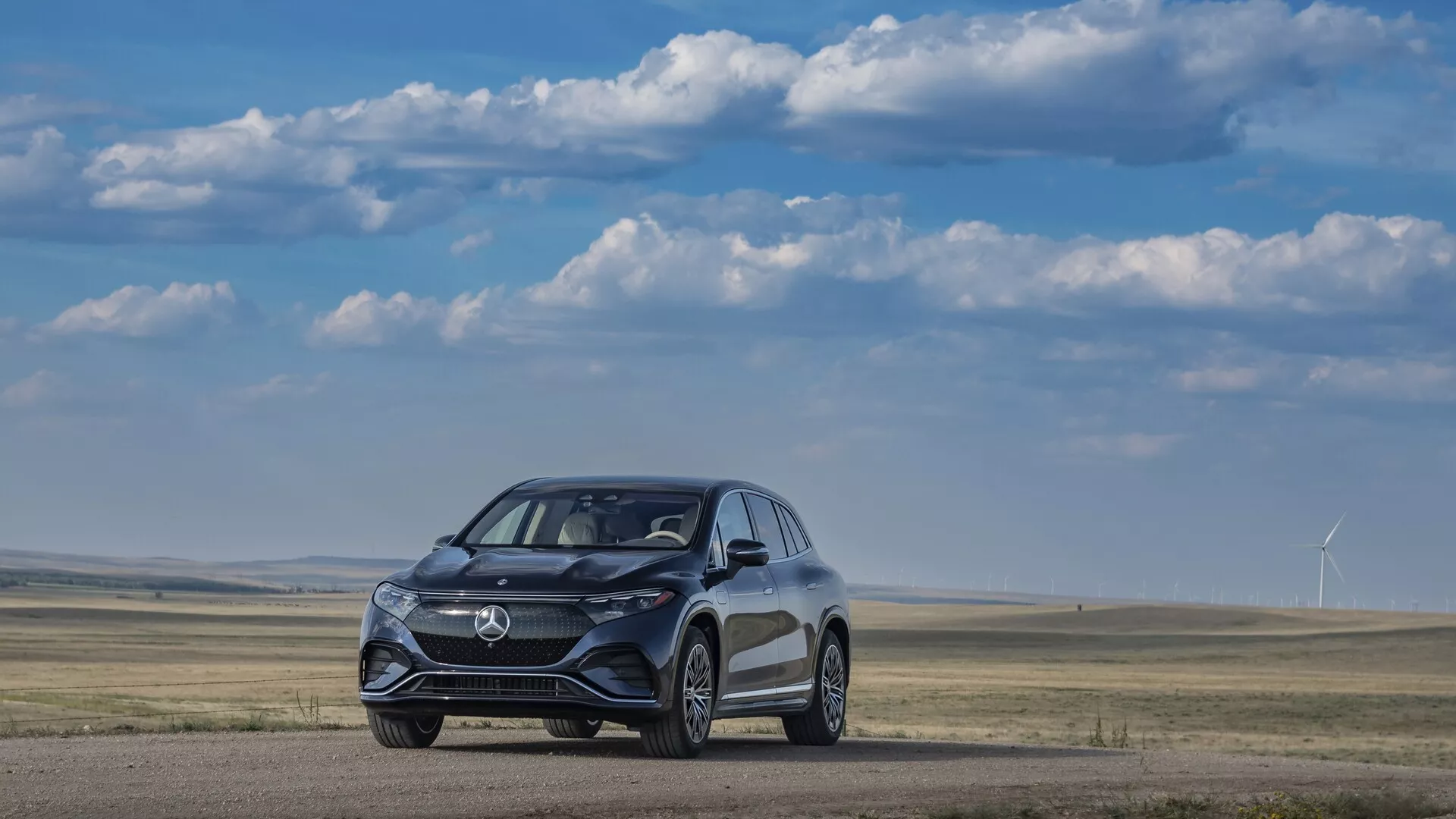 Slika prikazuje Mercedesa EQS SUV 2022 iz prednjega kota s treh četrtin, na ravni površini zemlje z travnikom in vetrnimi turbinami v ozadju pod jasno, delno oblačno nebo. Eleganten, aerodinamično oblikovan avto izstopa z moderno mrežico, LED žarometi in kolesi, osredotočenimi na zmogljivost. Vidne podrobnosti nakazujejo napredne sisteme za pomoč vozniku, vključno s senzorji in kamerami, integriranimi v prednji odbijač in stranska ogledala.