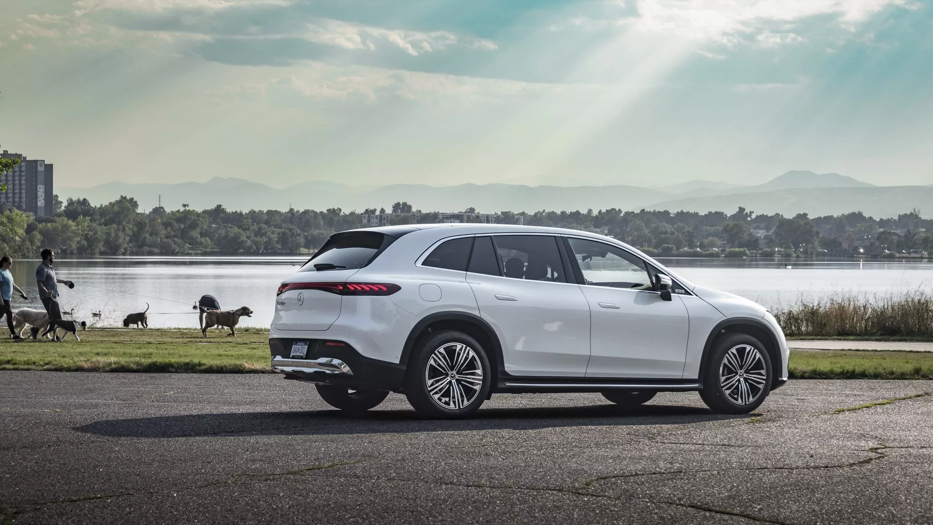 Profilna slika luksuznega Mercedes EQS SUV 2022, fotografiranega na asfaltirani cesti. SUV ima velika večkraka aluminijasta kolesa, elegantno linijo oken, ki se zožuje proti zadnjem delu s temnimi stekli, značilne aerodinamične linije karoserije, napredna stranska ogledala, streho, ki se proti koncu rahlo spušča z nosilcem in ovijalne LED zadnje luči. Vozilo je obkroženo s toplo svetlobo, kar poudarja njegov sodoben dizajn v mirnem okolju z vodnim telesom, drevesi in sprehajalci.