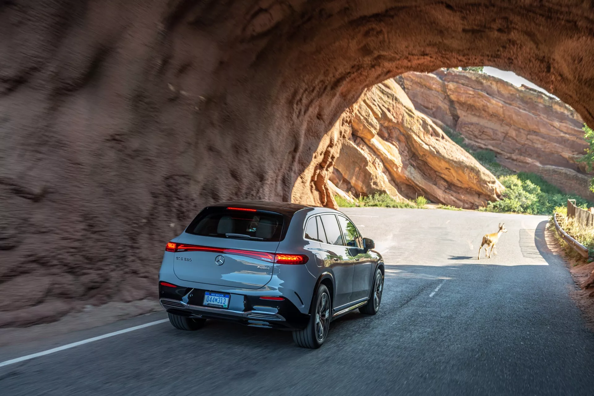 Zadnji in koten pogled na eleganten, modernen SUV, ki vozi skozi skalni tunel ob sončnem dnevu. Ključne značilnosti vključujejo vodoravne zavijalne zadnje luči, športni spojler, anteno v obliki morskega psa, odsevne okenske obrobe, dvojne izpušne cevi, petkraka aluminijasta platišča in na sredini nameščeno registrsko tablico pod zatemnjenim zadnjim oknom. Vozilo kaže napredno osvetljevalno tehnologijo in verjetno vključuje funkcije, kot so zadnja kamera in parkirni senzorji. Fotografija zajema gibanje in občutek raziskovanja na prostem z jelenom v ozadju.
