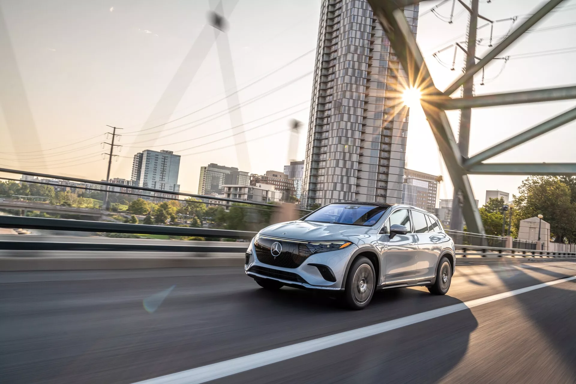 Dinamičen pogled na Mercedes EQS SUV 2022 v gibanju, zajet iz nizkega kota in iz sprednje stranske perspektive, prikazuje sprednjo rešetko, žaromete, pokrov motorja, vetrobransko steklo in del voznikove strani in strehe. Avto vozi po dvignjeni cesti, z varnostnimi ograjami in podporno strukturo vidno v ozadju. Slika poudarja sodobne oblikovalske elemente vozila, kot je izrazita sprednja rešetka, LED dnevne luči, aerodinamične linije na pokrovu motorja in strešno okno. Zamegljenost gibanja na kolesih in ozadju nakazuje zmerno do visoko hitrost, sončni odsevi pa nakazujejo zgodnje jutro ali pozno popoldne. Verjetno vključuje napredne sisteme za pomoč vozniku in prilagodljiv tempomat, zaradi česar je avto primeren za kategorije, kot so sodobna vozila, avtomobilska tehnologija, crossoverji, dinamika vozila in luksuzni avtomobili.