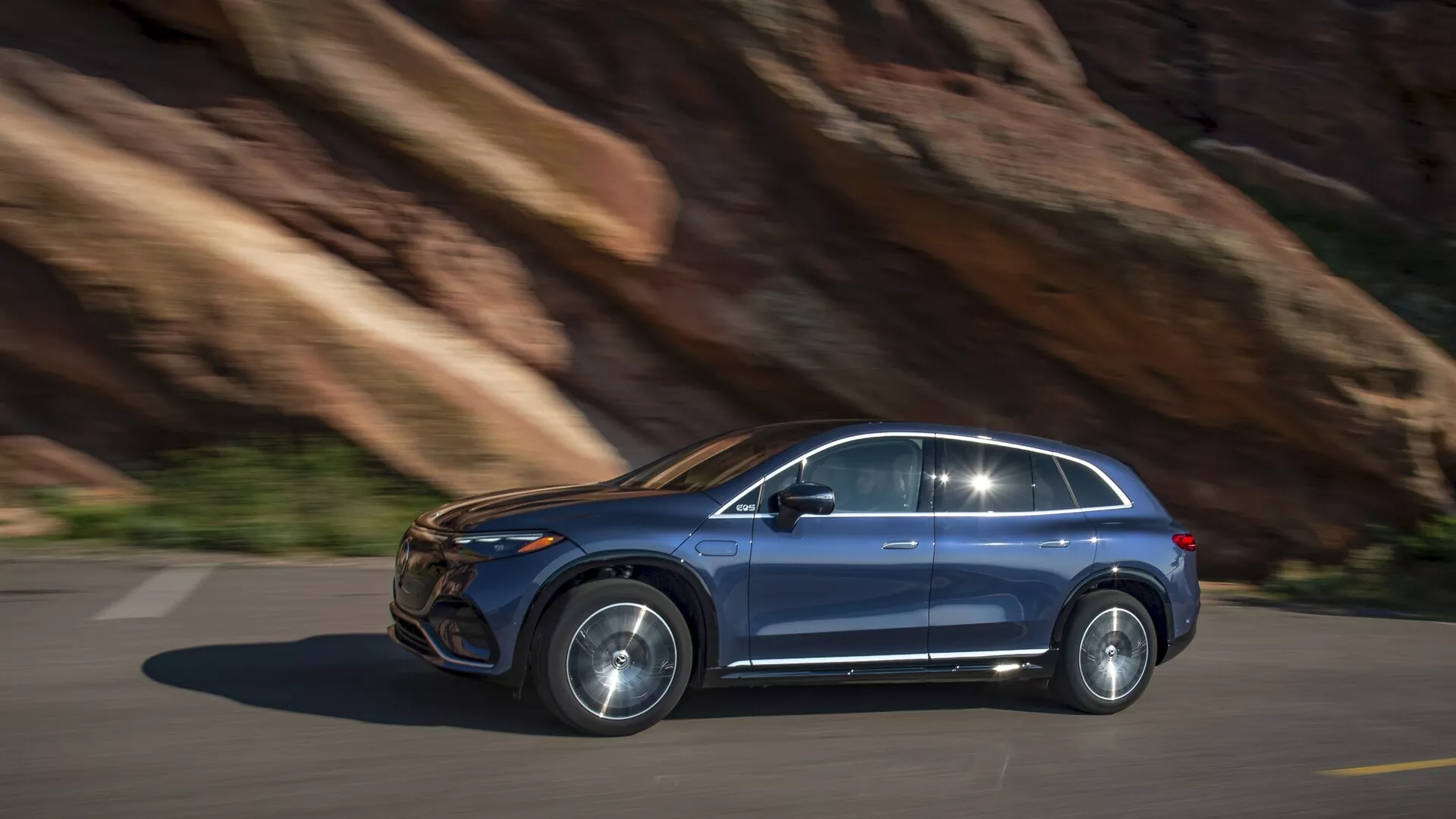 Mercedes EQS-SUV 2022 v gibanju, stranski profil. Premikanje z leve proti desni po asfaltirani cesti s kamnitim ozadjem, kar nakazuje na gorski teren. Desni stranski pogled prikazuje sodoben dizajn: vidna sprednja in zadnja vrata za potnike, dobro definirana stranska okna s oblogo, lita platišča in izrazit stranski ogledalnik. Kovinska barva, verjetno LED ali HID svetila, poravnane ročaji vrat. Možne napredne tehnološke funkcije: varnostni sistemi, info-zabava, električni/hibridni motor. Rahla meglitev gibanja kaže na dinamične zmogljivosti, dnevni posnetek z naravno svetlobo.