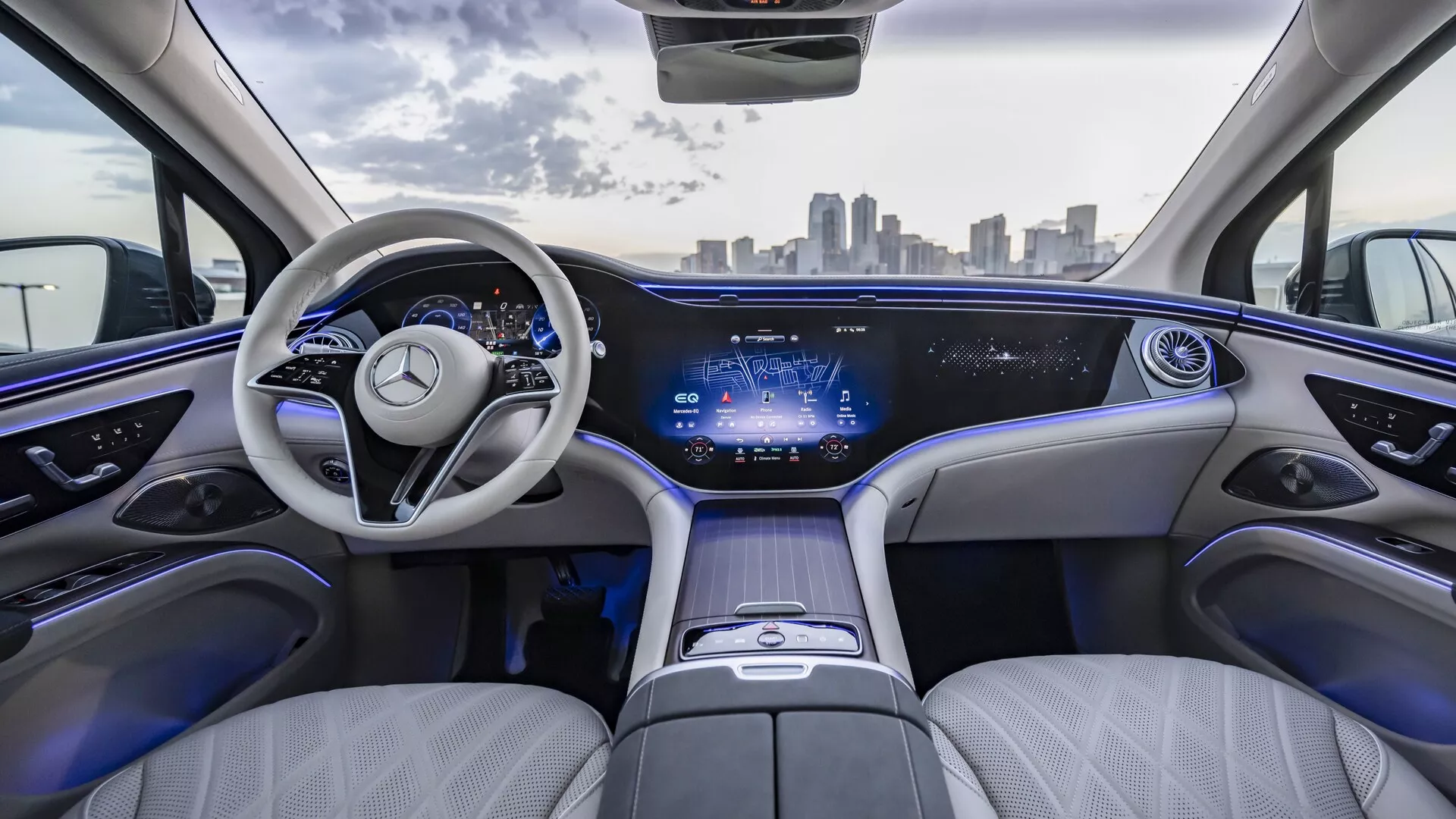 Notranji pogled na Mercedes EQS SUV 2022 iz zadnjih sedežev, z luksuzno svetlo usnjeno oblazinjenje, elegantno armaturno ploščo z neprekinjenim digitalnim prikazom, sodobnim dvokrakim volanom z emblemi Mercedes-Benz, prostorno in udobno postavitvijo sedežev, ambientalna osvetlitev, sredinska konzola z dotikajočimi se gumbi in panoramska streha. Urban obzorje vidno skozi vetrobransko steklo.