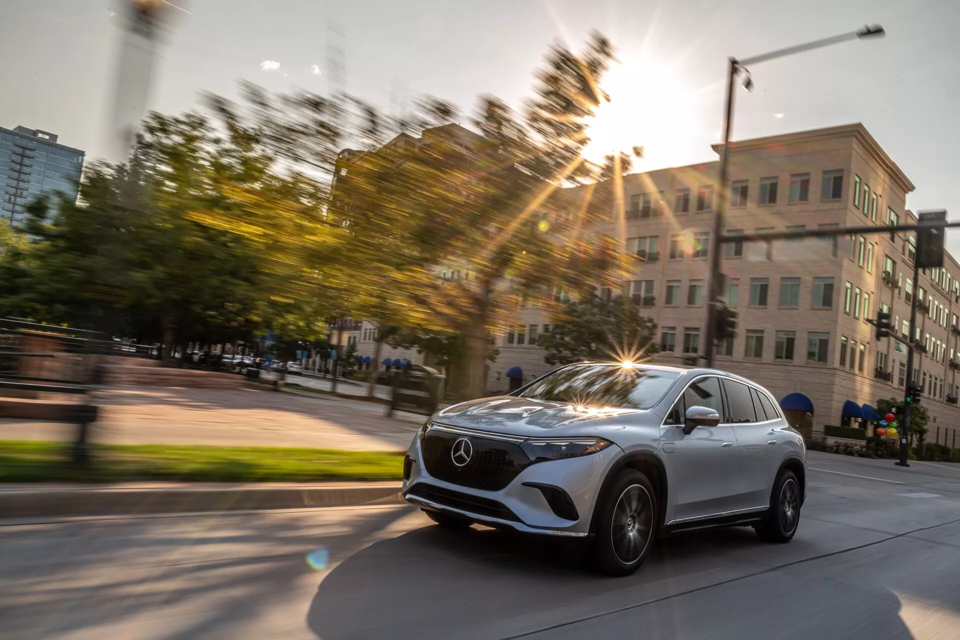 Dinamična slika Mercedes EQS SUV 2022, posneta v gibanju iz rahlo diagonalnega prednjega tričetrtinskega pogleda. Fotografija poudarja sprednjo mrežo vozila z Mercedesovim emblemom, elegantne LED žaromete, kolo in blatnik na voznikovi strani ter del vetrobranskega stekla in voznikova vrata. Kolesa avtomobila so nekoliko obrnjena v desno, kar nakazuje na zavijanje ali spremembo smeri. Sončna svetloba ustvarja učinek leče, ki dodaja energijo fotografiji, posneti v urbanem okolju z zgradbami in zelenjem, med zlatimi urami. Zameglitev gibanja v ozadju poudarja hitrost in gibanje.