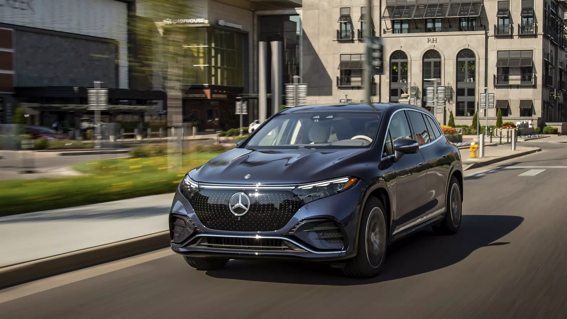 Dinamičen posnetek Mercedes EQS SUV 2022 v gibanju na ulici, posnet iz nizke treh četrtinske perspektive s strani voznika. Luksuzno vozilo poudarja svojo izrazito sprednjo rešetko z velikim emblemom, elegantne LED žaromete z dnevnimi lučmi in aerodinamično zasnovo. Aluminijasta platišča in stranska ogledala v barvi karoserije z vgrajenimi smerniki poudarjajo razkošje. Mestno ozadje z efektnim zamegljenem gibanjem poudarja napredne tehnološke značilnosti avtomobila in njegovo prisotnost na cesti.
