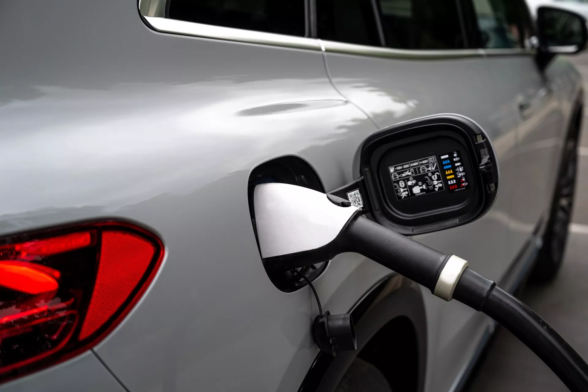 Poudarek na območju priključka za polnjenje na strani srebrnega Mercedes EQS-SUV 2022, s poudarkom na priključenem polnilnem kablu, odprtem priključku, ki razkriva digitalni zaslon, ki označuje stanje polnjenja, z rahlo zamegljenim ozadjem, ki prikazuje dele zadnje luči in odsev drugega vozila.