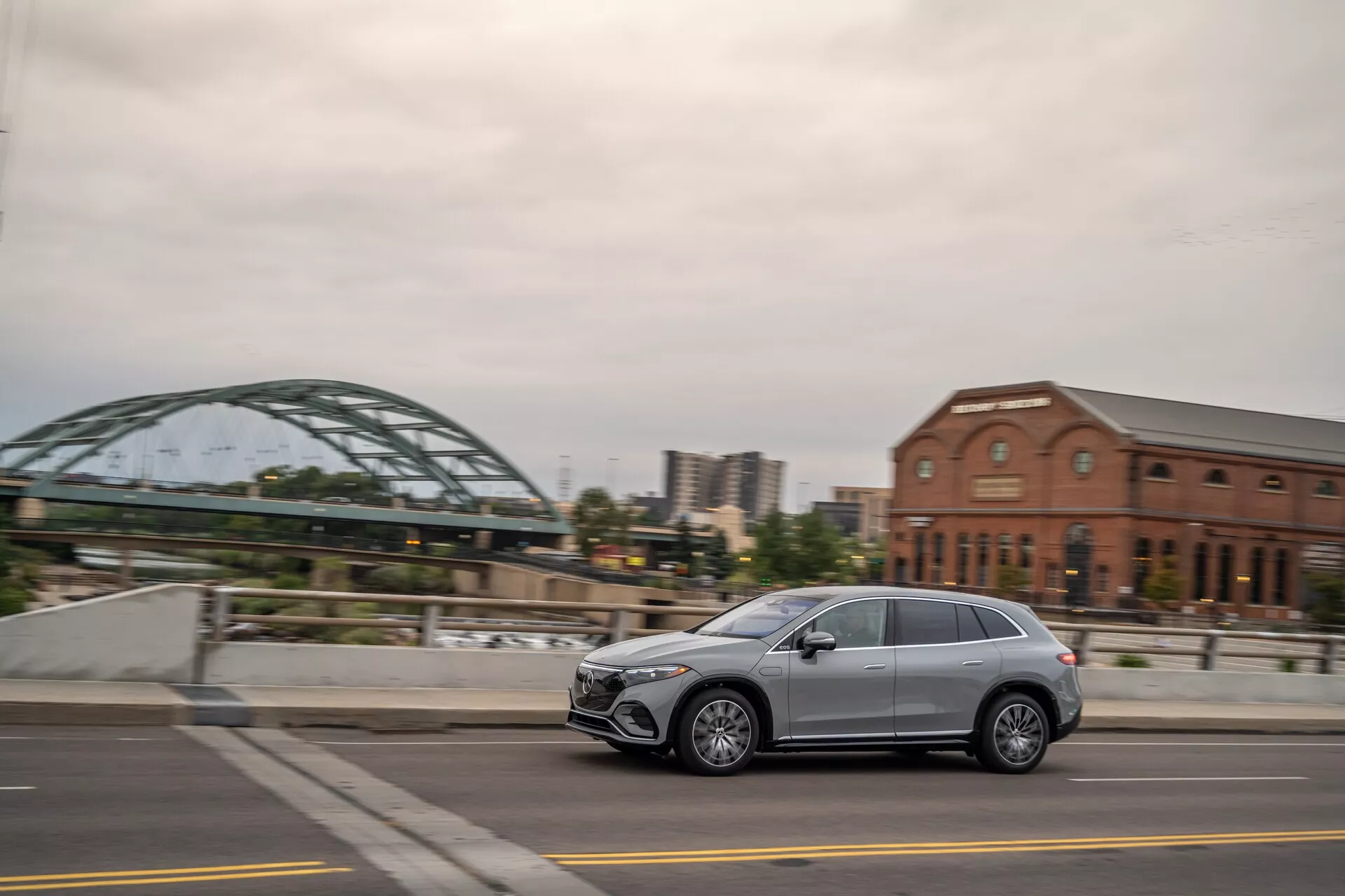 Stranski profil sodobnega Mercedesa EQS-SUV 2022, ki vozi po mestni ulici, ki prikazuje eleganten dizajn, pet vrat, lita platišča in LED žaromete. Posnet iz rahlo zadnjega kota z zamegljenim ozadjem.
