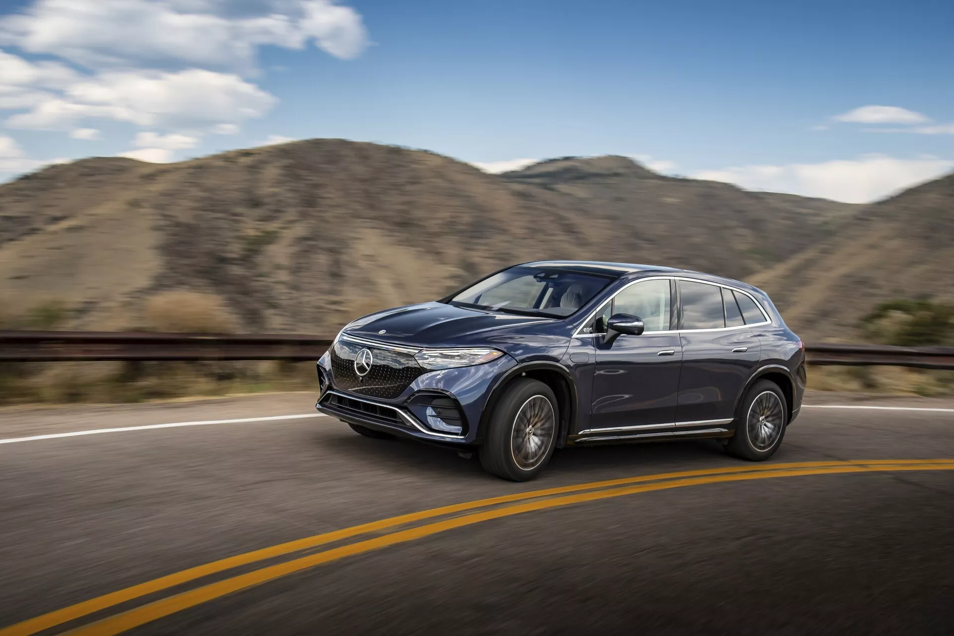 Gibajoča se fotografija modernega Mercedesa EQS-SUV 2022, ki vozi po gladki asfaltni cesti z rumeno ločilno črto pod jasnim nebom. Avto je viden iz rahlega sprednjega stranskega kota, poudarja voznikovo stran, vključno s sprednjo masko, zasnovo žarometov, voznikovimi vrati, stranskim ogledalom in delom vetrobranskega stekla. Vozilo je v ospredju s zamegljenim ozadjem hribov, kar nakazuje gibanje. Vidne so oblikovne značilnosti, kot so aluminijasta platišča, izklesani stranski paneli, značilne stilske linije in potencialna tehnologija ADAS.