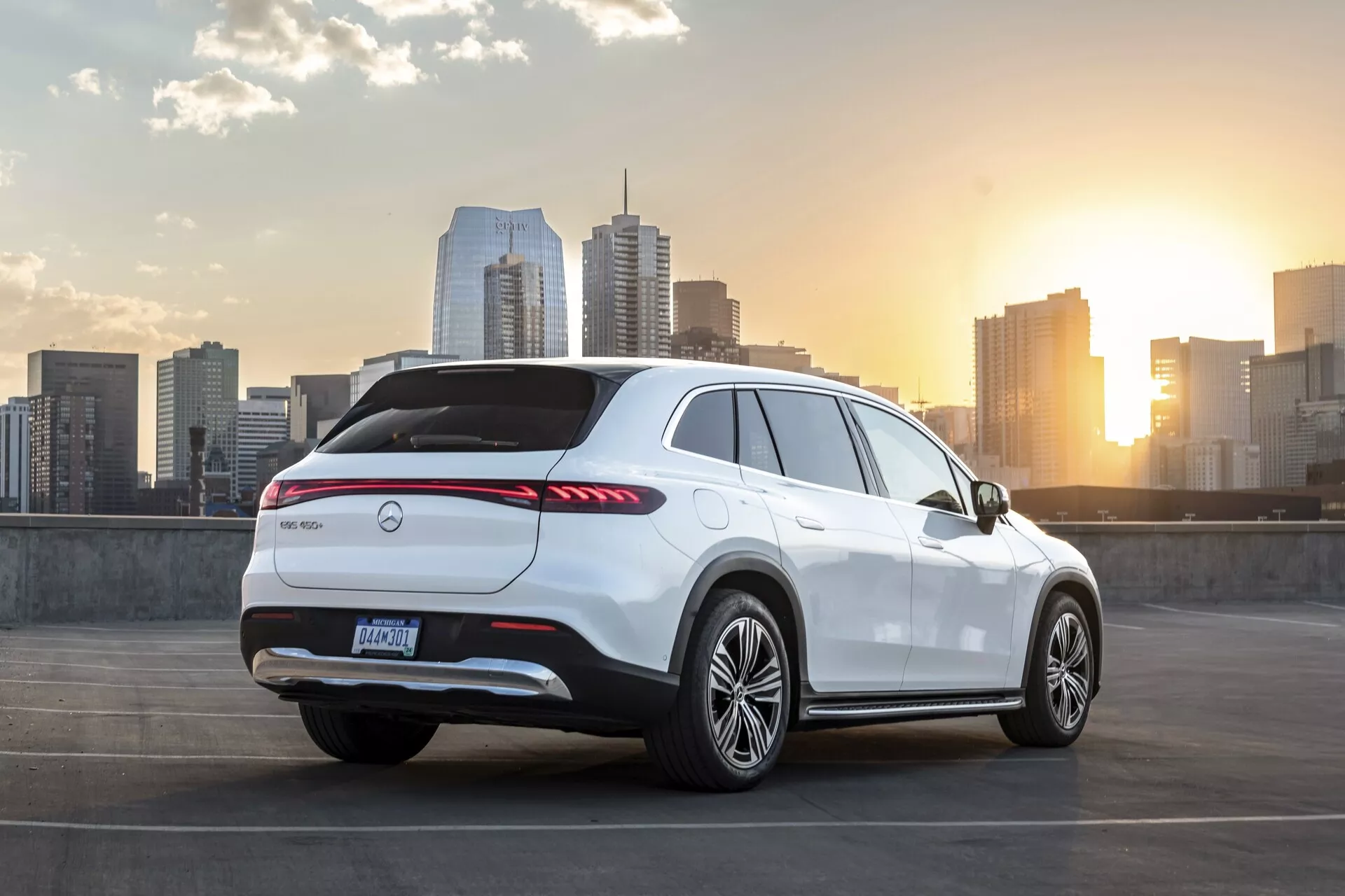 Slika belega Mercedes EQS-SUV 2022 parkiranega na zgornji ravni parkirne strukture. Vozilo je vidno iz treh četrt zadnjega kota, prikazuje zadnji del, stransko stran potnika in del strešne linije. Vključuje podaljšane zadnje LED luči, proizvajalčev emblem na sredini zadnjih vrat, zadnji difuzor, zadnji spojler, aluminijasta platišča in visoko linijo pasu s športno nagnjeno streho. Scena vključuje mestno obzorje in sončni zahod v ozadju, poudarjajoč tematiko urbanega razkošja in sodobnega življenjskega sloga.