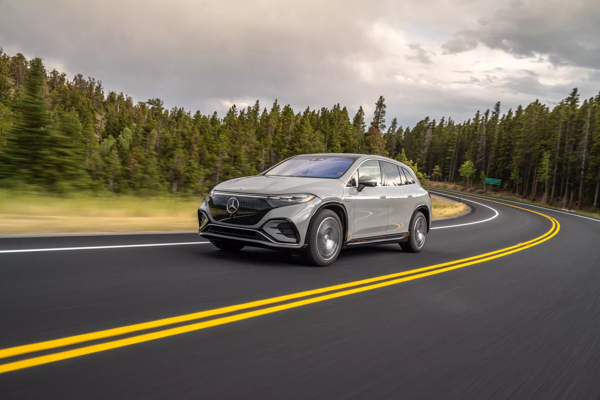 Ta slika prikazuje Mercedes-Benz EQS SUV 2022 v gibanju iz treh četrtin spredaj z vidnim čelnim dizajnom in stranskim profilom. Vozilo je pozicionirano na levi, vozi po dobro vzdrževani asfaltni cesti z dvojnimi rumenimi črtami in zavojem v desno. Rahlo zamegljenost koles in rahlo zamegljenost zaradi počasnejše hitrosti zaklopa poudarja hitrost in dinamično gibanje. Oblakane svetlobne razmere povzročajo mehke sence in enakomerno osvetlitev vozila. Vidne so tehnološke funkcije, kot so LED žarometi, dnevne luči, velika prednja maska z emblemi in aerodinamična stranska ogledala z morebitnimi senzorji. Srebrna ali svetlo siva barva kontrastira z bleščečimi črnimi podrobnostmi, kot so spodnja maska, obloge kolesnih lokov in okenski okviri, poudarjajo elegantne linije, ukrivljene blatnike in strešno linijo, podobno kupeju. Primeren za avtomobilske, tehnološke, gibljive fotografije in luksuzna vozila.
