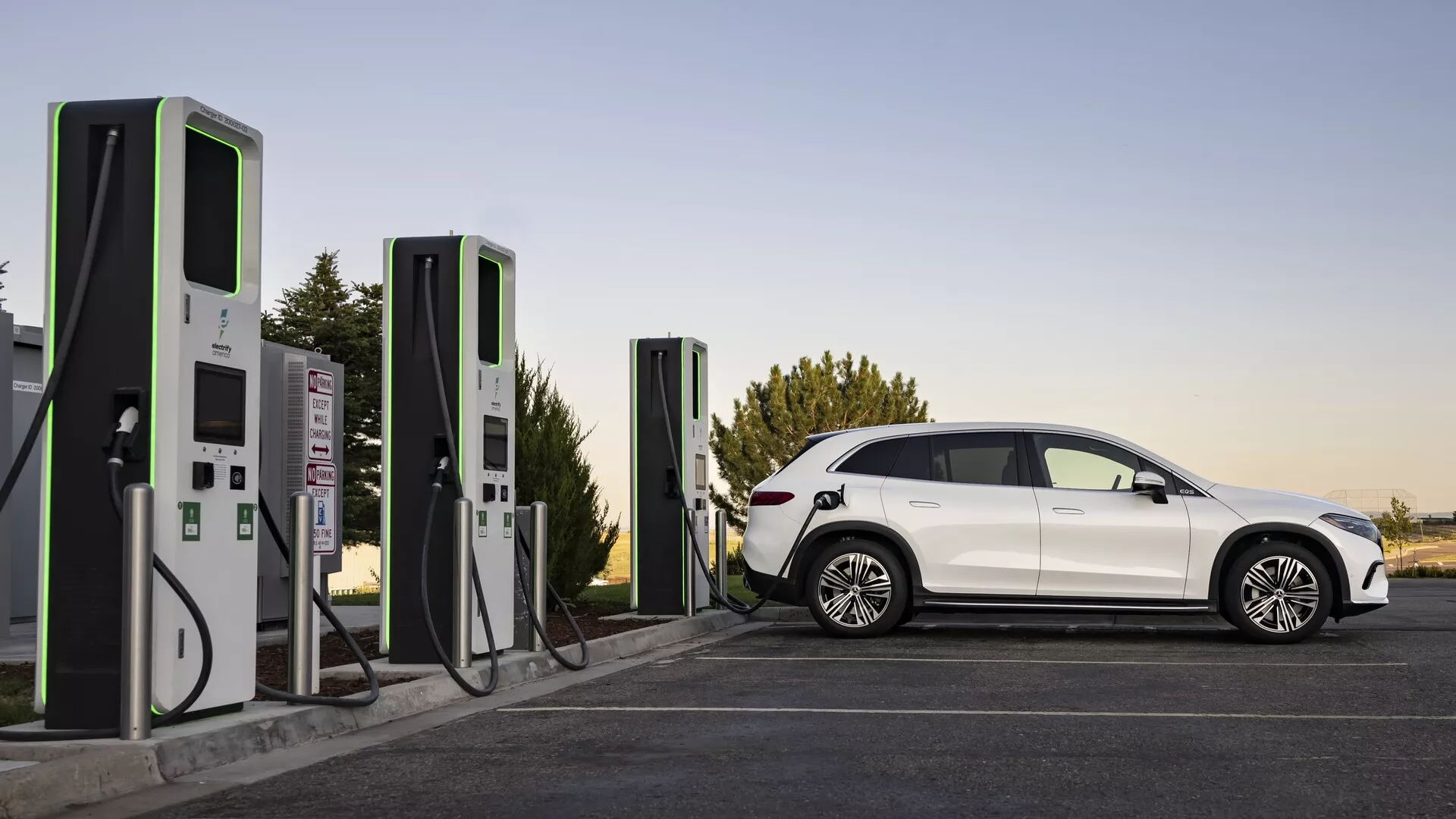 Eleganten in sodoben električni avtomobil (EV), Mercedes EQS-SUV 2022, predstavljen na polnilni postaji v mehki svetlobi, bodisi pozno popoldne ali zgodaj zjutraj. Celoten stranski profil razkriva aerodinamične linije, značilna kolesa, zatemnjena okna in jasno definiran potniški prostor. Vozilo je priključeno na sodobno polnilno postajo, kar poudarja osredotočenost na trajnostni prevoz in infrastrukturo za električna vozila.