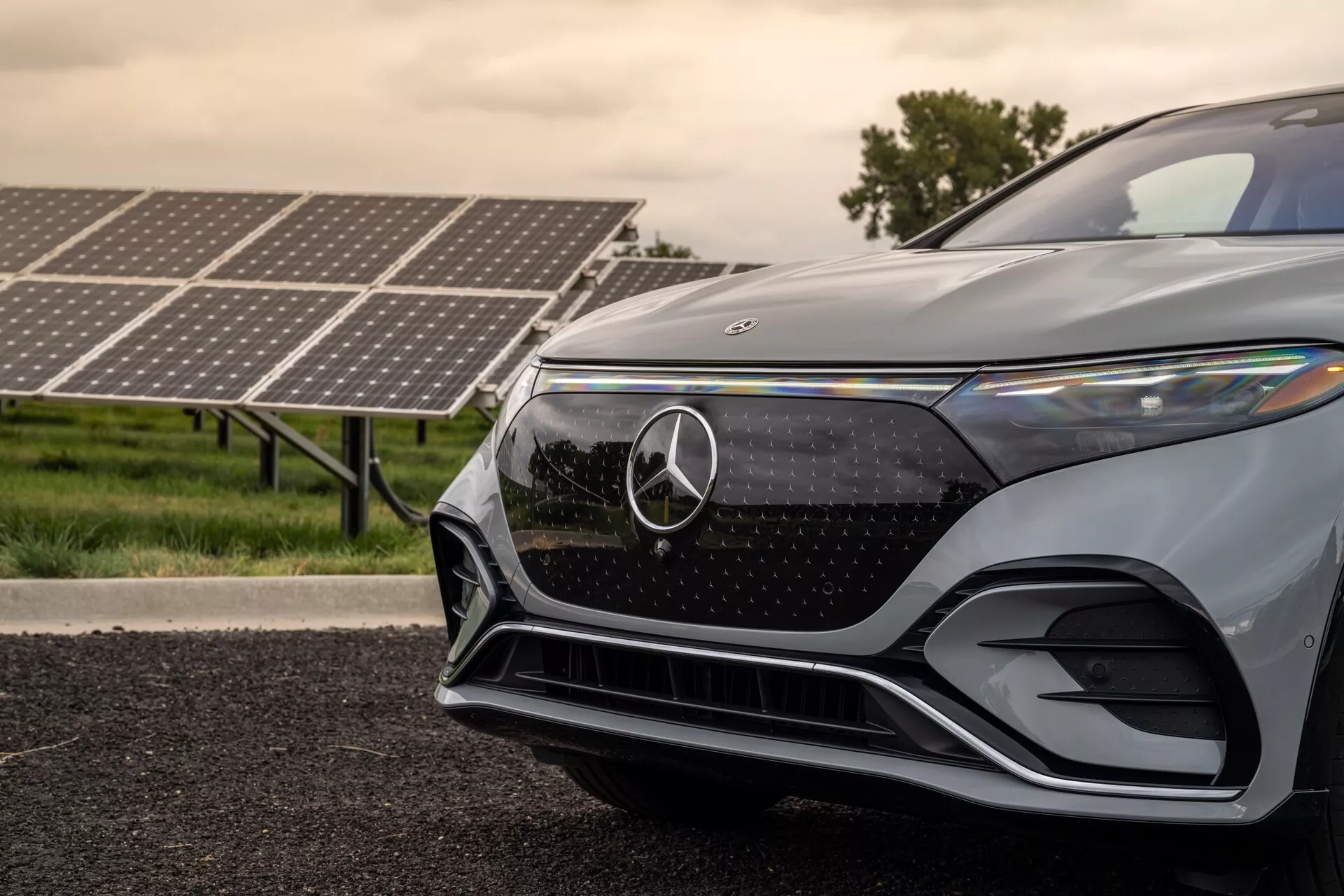 Od blizu sprednjega levega četrt dela sodobnega Mercedes-Benz EQS SUV 2022, posnetega iz nizkega kota. Slika poudarja rešetko z Mercedes-Benz emblemom, elegantni kotni LED žaromet, aerodinamični sprednji odbijač in kovinsko lakiranje. Sončni kolektorji v ozadju namigujejo na električno ali hibridno naravo avtomobila, kar poudarja njegove napredne in okolju prijazne lastnosti.