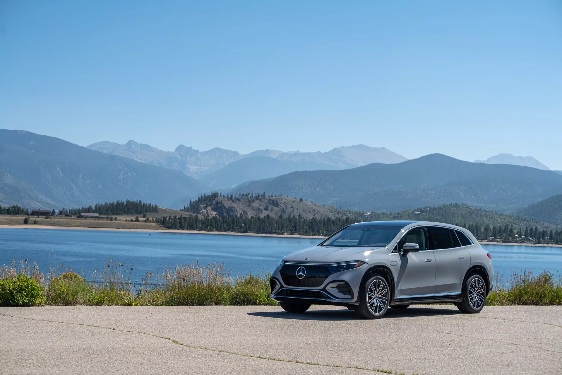 Slika prikazuje sodoben Mercedes EQS SUV 2022, parkiran na tlakovanem območju s slikovitim ozadjem vode in gorskih verig, posnet z nizkega kota, da poudari njegov postavo in oblikovne linije. Avto je viden iz stranskega profila z vidnim sprednjim žarom, kotnimi LED žarometi, velikimi aluminijastimi kolesi, aerodinamičnimi stranskimi ogledali in kovinsko barvo.