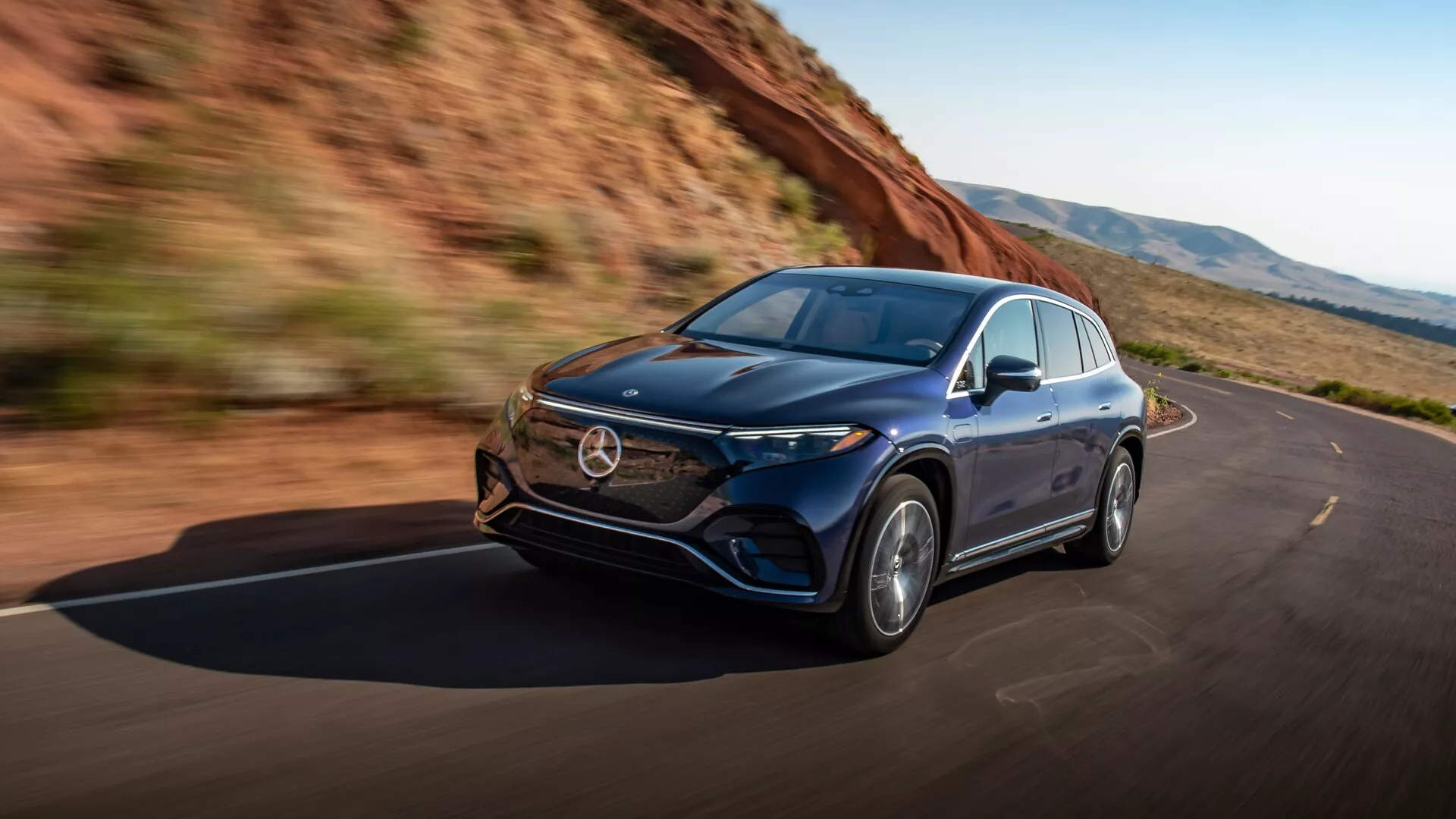 Dinamičen posnetek Mercedes EQS-SUV 2022, ujet iz kot spredaj levo pod kotom tri četrt, ki prikazuje vozilo v gibanju. Zajema elegantna leva stran avtomobila, prednja hauba, žarometi z napredno LED tehnologijo, rešetko, prednji odbijač in lita platišča. Aerodinamična oblika karoserije nakazuje na luksuz in napredno inženirstvo, izpostavlja zmogljivost in vozniško izkušnjo. Posnetek oddaja občutek svobode in zmogljivosti na čistih cestah, obdanih z naravo.