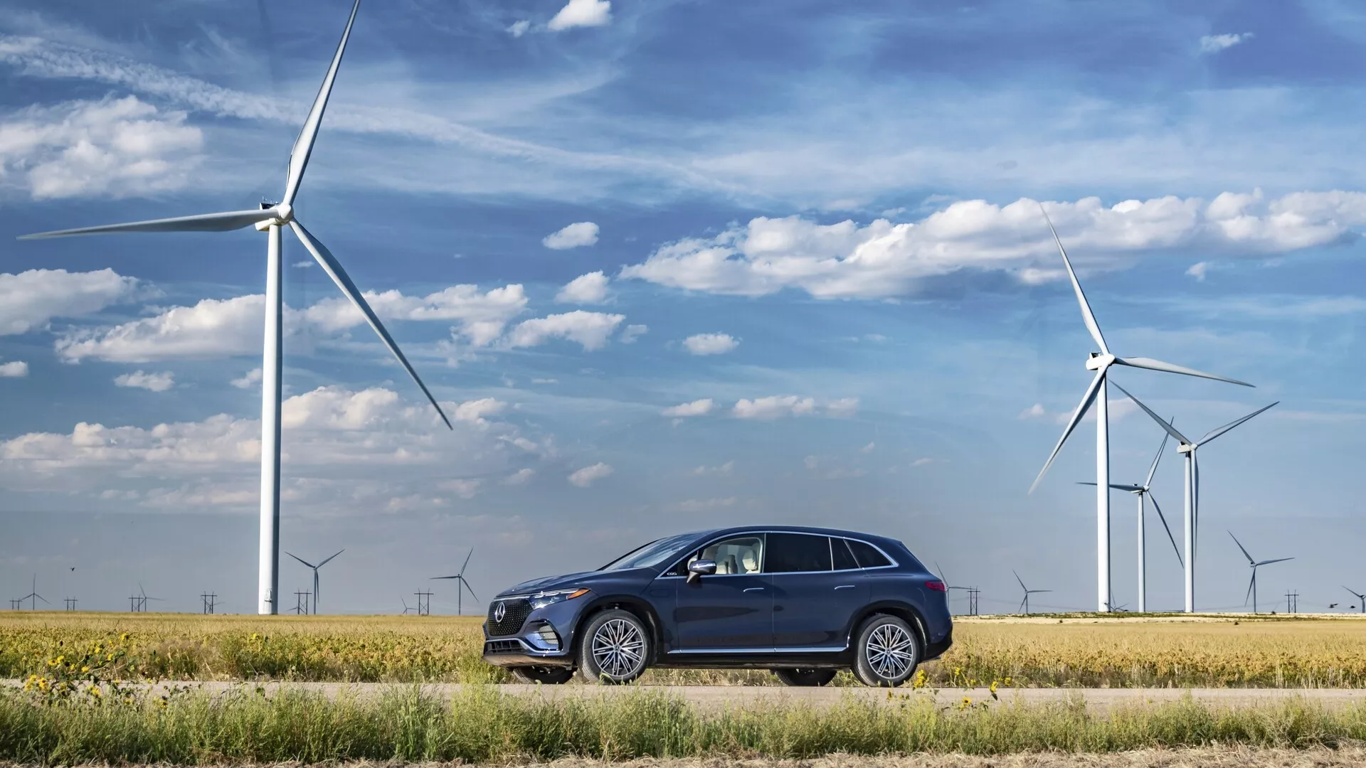 Mercedes EQS-SUV 2022 parkiran v okolju prijazni krajini z vetrnimi turbinami v ozadju, ki prikazuje sodoben, eleganten dizajn s temnim barvnim ohišjem, večkrakimi aluminijastimi platišči, vgrajenimi ročaji vrat, kromiranimi obrobami oken, kotnimi LED žarometi in vgrajenimi smerniki v ogledalih.