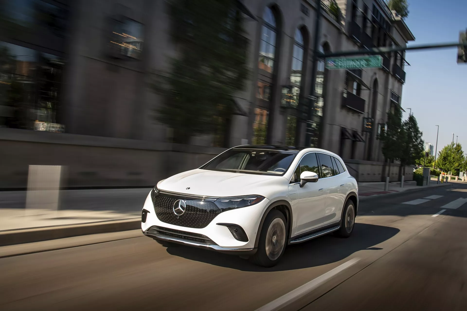 Slika prikazuje Mercedes EQS-SUV 2022 v gibanju na mestni ulici iz trikotnega sprednjega kota, ki poudarja njegovo sprednjo masko, oblikovano pokrov motorja, elegantne kotne LED žaromete in del zasnove koles. Bela barva vozila kontrastira z zamegljenim mestnim ozadjem, kar poudarja njegovo hitrost in dinamično prisotnost. Velik emblem na rešetki, aerodinamično oblikovano bočno ogledalo in napredna konfiguracija žarometov nakazujejo njegove moderne, luksuzne in tehnološko napredne značilnosti.