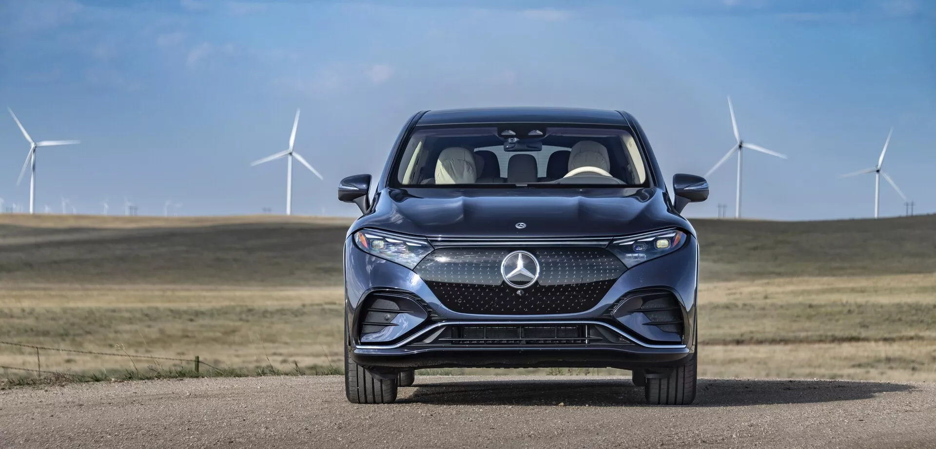 Sprednji pogled Mercedes EQS-SUV 2022 proti ozadju jasnega neba in vetrnih turbin. Vidna mreža vozila vključuje velik stiliziran emblem s kompleksnimi vzorci, ki verjetno vsebujejo senzorje za sisteme asistence voznikom. Vidni so elegantni horizontalni LED žarometi z integriranimi dnevnimi svetili. Športni sprednji odbijač vključuje velike zračne reže, konture pokrova motorja pa se nagibajo naprej za izboljšano aerodinamiko. Delni pogled na sprednjo četrtino razkriva del blatnikov, kar je značilno za premium dizajn. Odsev vetrobranskega stekla in delno vidno stransko ogledalo potnika nakazuje napredne funkcije, kot so smerokazi in nadzor mrtvih kotov. Napredni sistemi asistence vozniku in znaki trajnosti se nakazujejo skozi okolje z vetrnimi turbinami.
