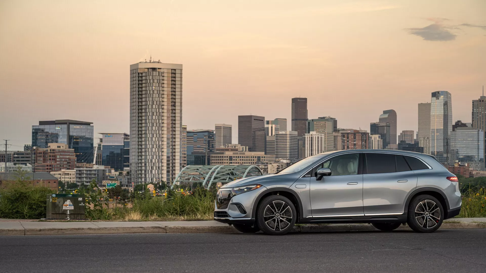 Stranski pogled na sodoben srednje velik Mercedes EQS SUV 2022, parkiran ob mestni silhueti ob mraku, prikazuje eleganten aerodinamičen dizajn, aluminijasta platišča, strešne nosilce, možno strešno okno in napredne funkcije, kot so integrirani smerniki v vzvratnih ogledalih in brezključni vstop.