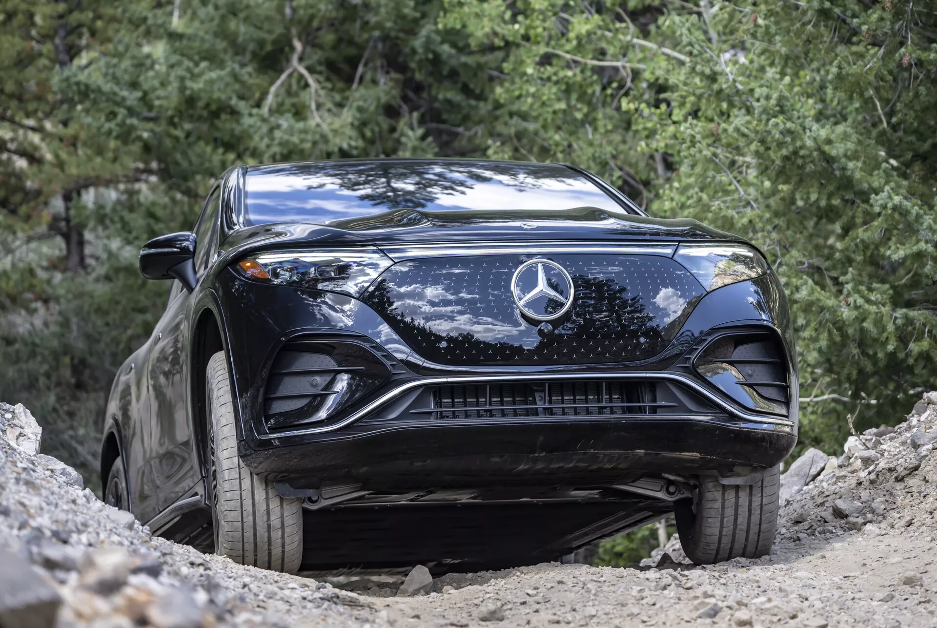 Slika iz spodnjega sprednjega kota črnega Mercedes-Benz EQS SUV 2022, ki poudarja oblikovan pokrov motorja, velik sredinski zvezdasti emblem, zapleteno mrežo, elegantne LED žaromete in spodnje dovode zraka. Zajeto v naravnem zunanjem okolju, vozilo, ki se nahaja na naklonu, prikazuje večjo oddaljenost od tal in terenske zmogljivosti. Vozilo je čisto z nekaj umazanije, kar poudarja lahko uporabo izven ceste in napreden avtomobilski dizajn.