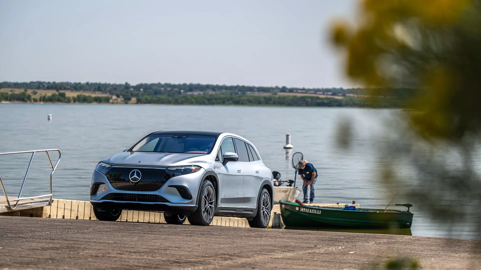 Slika prikazuje razkošno vozilo Mercedes-Benz EQS SUV 2022, parkirano ob jezeru ali rečnem pomolu. Vozilo je rahlo postavljeno pod kotom, kar omogoča pogled na tri četrtine spredaj in poudarja značilno masko, natančne LED žaromete, sprednji odbijač, vetrobransko steklo in del zadnjega dela. Okolje je mirno, z vodno površino in drugimi plovili v ozadju, kar nakazuje rekreacijski kontekst. Scena je dobro osvetljena, kar poudarja elegantne oblikovne linije in obrise vozila, medtem ko zamegljeni listi ustvarjajo naravni okvir.