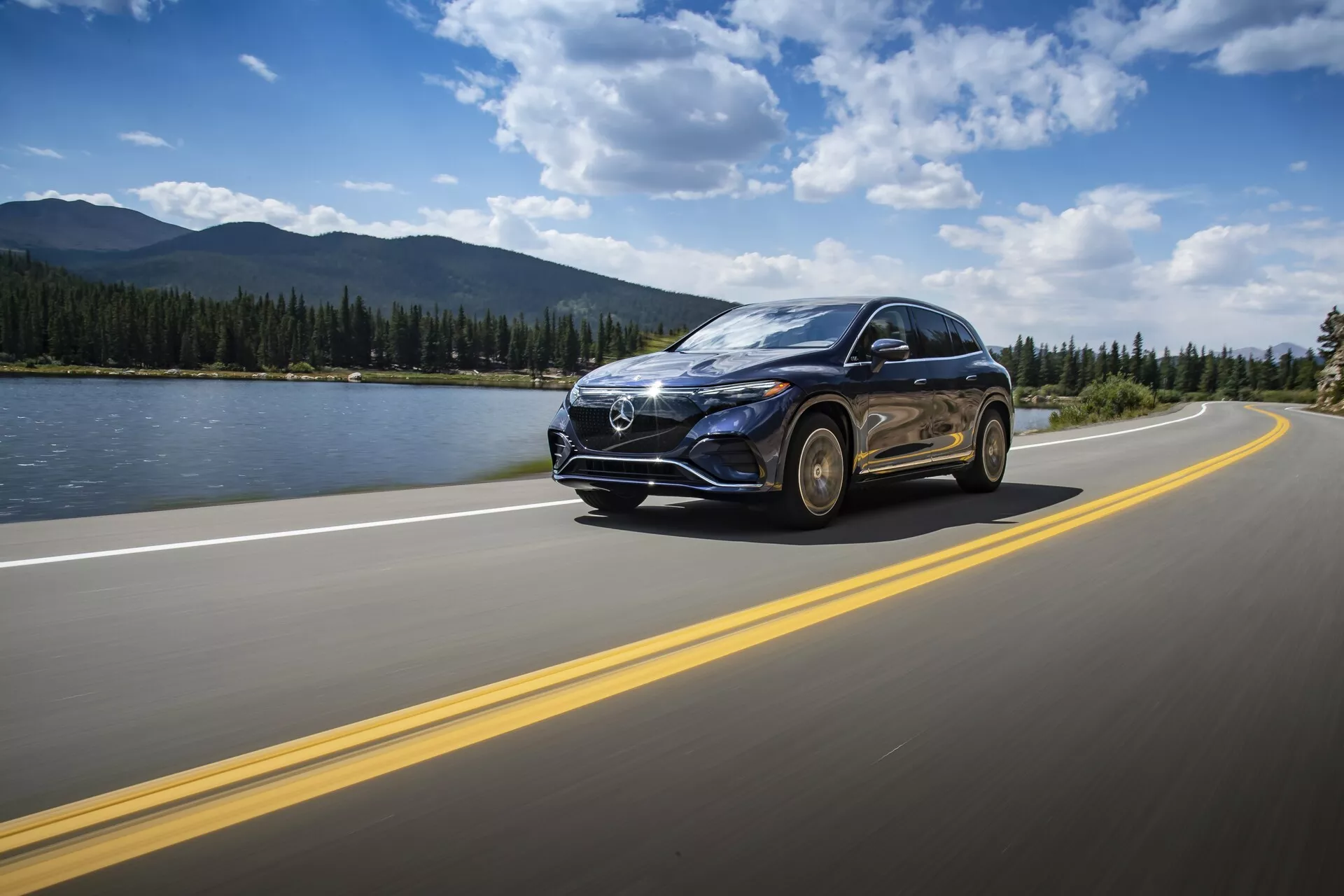 Mercedes EQS SUV 2022 vozi po asfaltirani cesti z dvojnimi rumenimi črtami pod delno oblačnim dnevnim nebom. Vozilo je zajeto iz nizkega sprednjega tričetrtinskega zornega kota, poudarjajoč izrazito sprednjo rešetko z velikim emblemom, LED žaromete, elegantne linije pokrova motorja, platišča iz lahke zlitine in stransko ogledalo z morebitnimi vgrajenimi smerniki. Ozadje vsebuje miren pokrajinski prizor z vodnim telesom, gorami in čistim obzorjem, poudarjajoč občutek potovanja in avanture. Premikanje avta je poudarjeno z rahlo zameglitvijo gibanja.