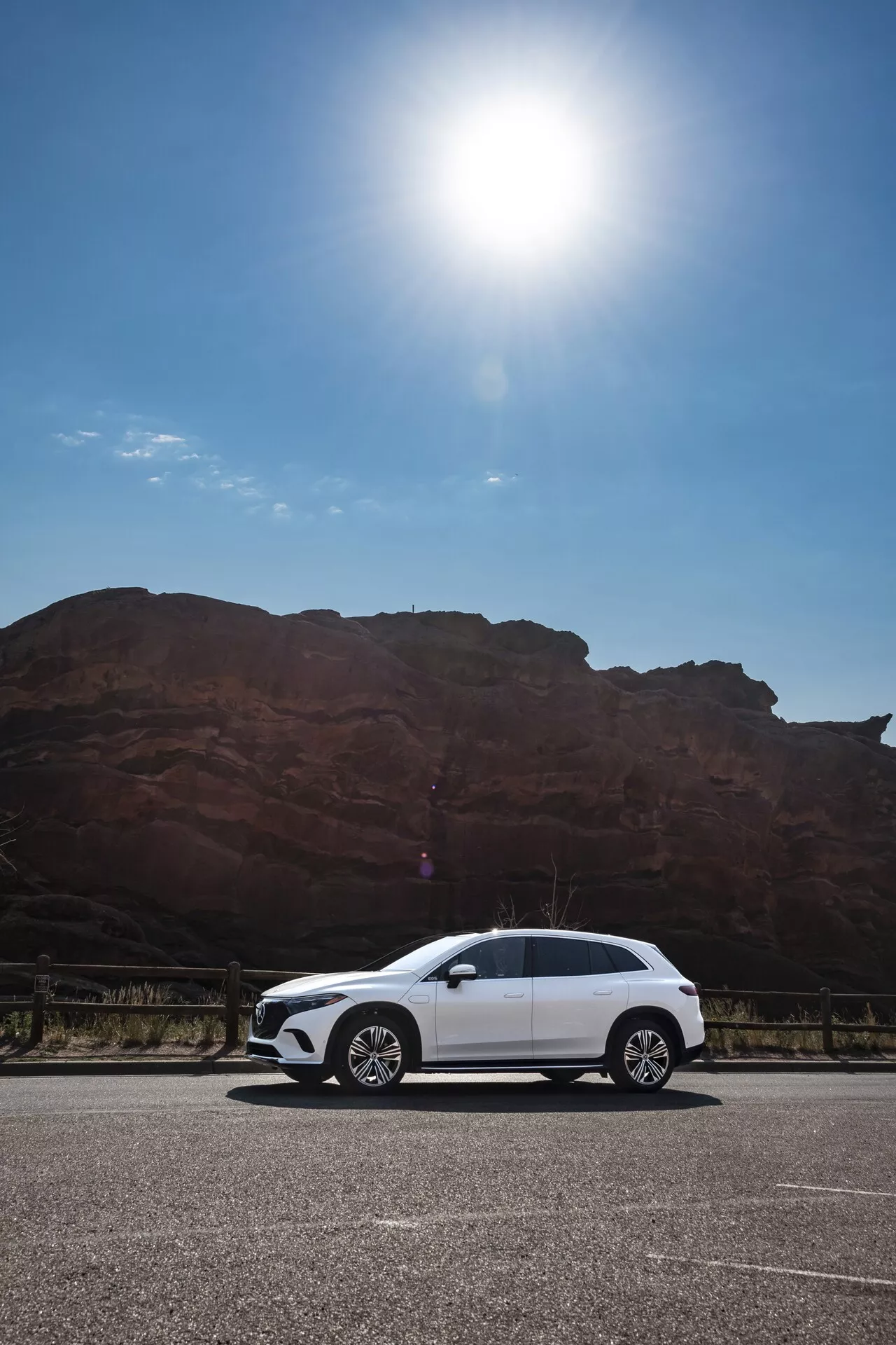 Dnevna slika belega Mercedesa EQS SUV 2022, parkiranega na tlakovanem območju pred veliko, rdečkasto-rjavo skalno formacijo, posneta pod nizkim kotom, ki prikazuje stranski profil avtomobila, vključno z ročaji vrat, stranskimi ogledali, platišči in obrisi karoserije pod jasnim nebom z bleščanjem sonca v zgornjem levem kotu.