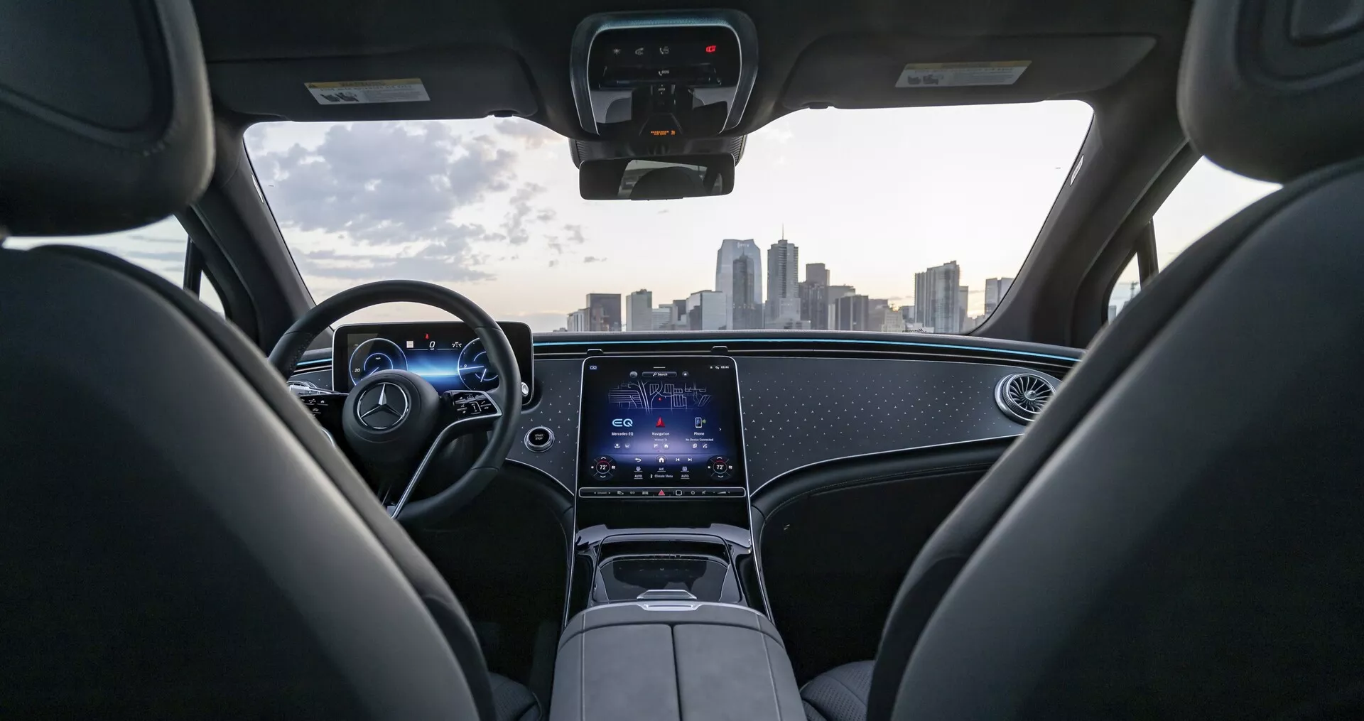 Pogled na notranjost Mercedes EQS-SUV 2022 iz perspektive zadnjega sedeža, prikazovanje modernega armaturne plošče z ambientno osvetlitvijo, digitalnih instrumentov in usnjenega volana z vgrajenimi kontrolami. Centralno konzolo prevladuje velik zaslon na dotik za infotainment in povezovalne funkcije. Veternik ponuja jasen in neoviran pogled na obzorje mesta, kar poudarja napredno tehnologijo in luksuzni dizajn avtomobila.