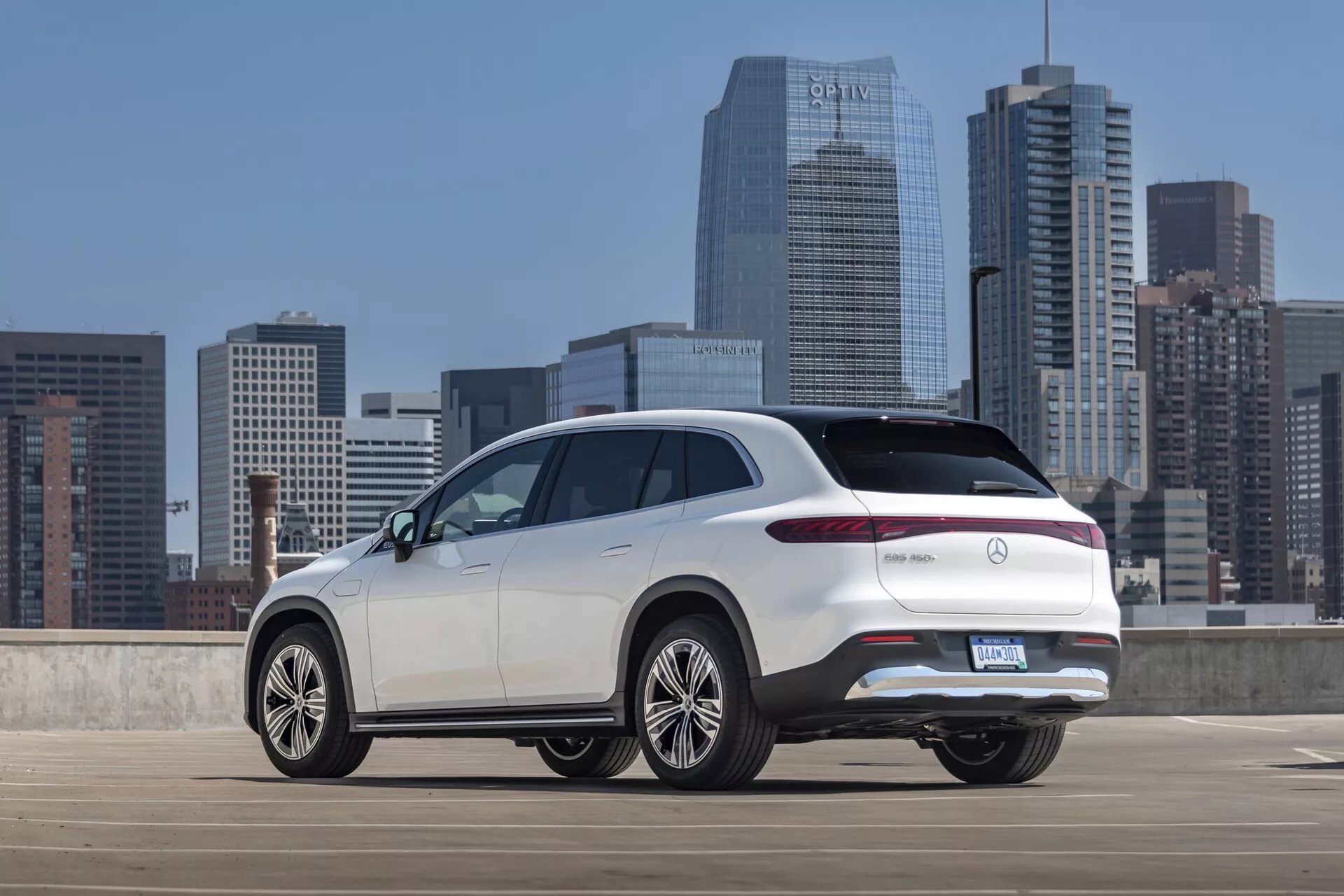 Tritrtninski zadnji pogled na bel Mercedes EQS-SUV 2022 iz rahlo dvignjenega kota, ki prikazuje zgornji del zadnjega dela, stranski profil in izrazite oblikovne značilnosti, kot so elegantne LED zadnje luči, zadnja vrata, zadnji odbijač in velika večkraka platišča iz lahke zlitine. SUV je parkiran v odprti mestni parkirni strukturi z mestnim obzorjem v ozadju, kar poudarja njegovo moderno, vrhunsko obliko z morebitnimi naprednimi funkcijami, značilnimi za luksuzne modele.