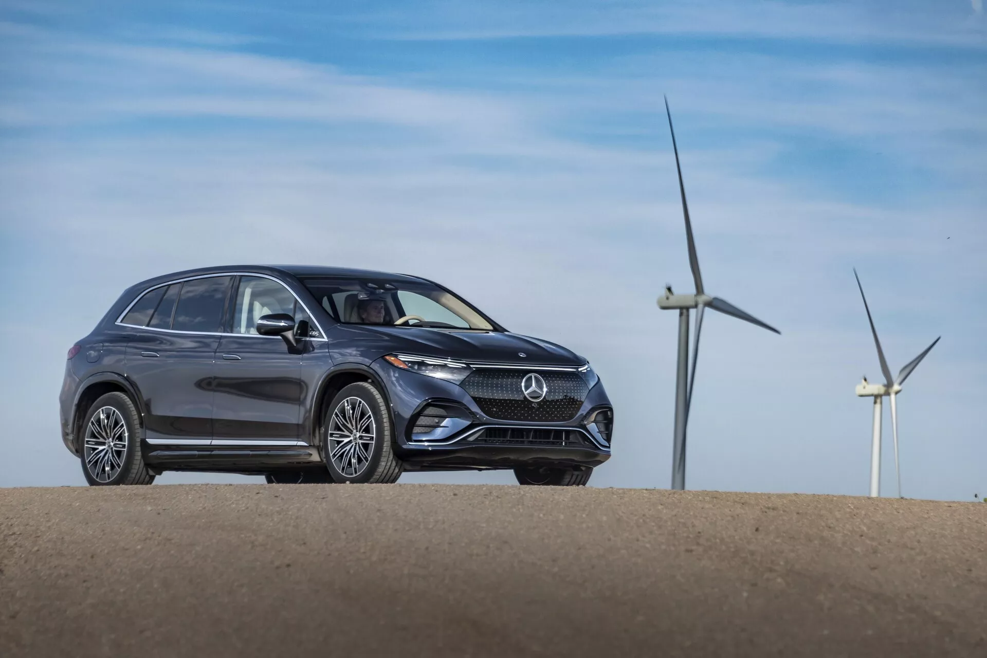 Trikrat položni prednji pogled na siv Mercedes EQS-SUV 2022, postavljen na majhnem hribu, ki prikazuje sodoben dizajn z elegantnimi linijami karoserije, značilne oblike žarometov, izrazito sprednjo masko z logotipom, stacionarnimi kolesi in platišči iz lahke zlitine. Veterne turbine v ozadju nakazujejo na vetrno farmo ali podeželsko območje, kar morda nakazuje na ekološke lastnosti vozila.
