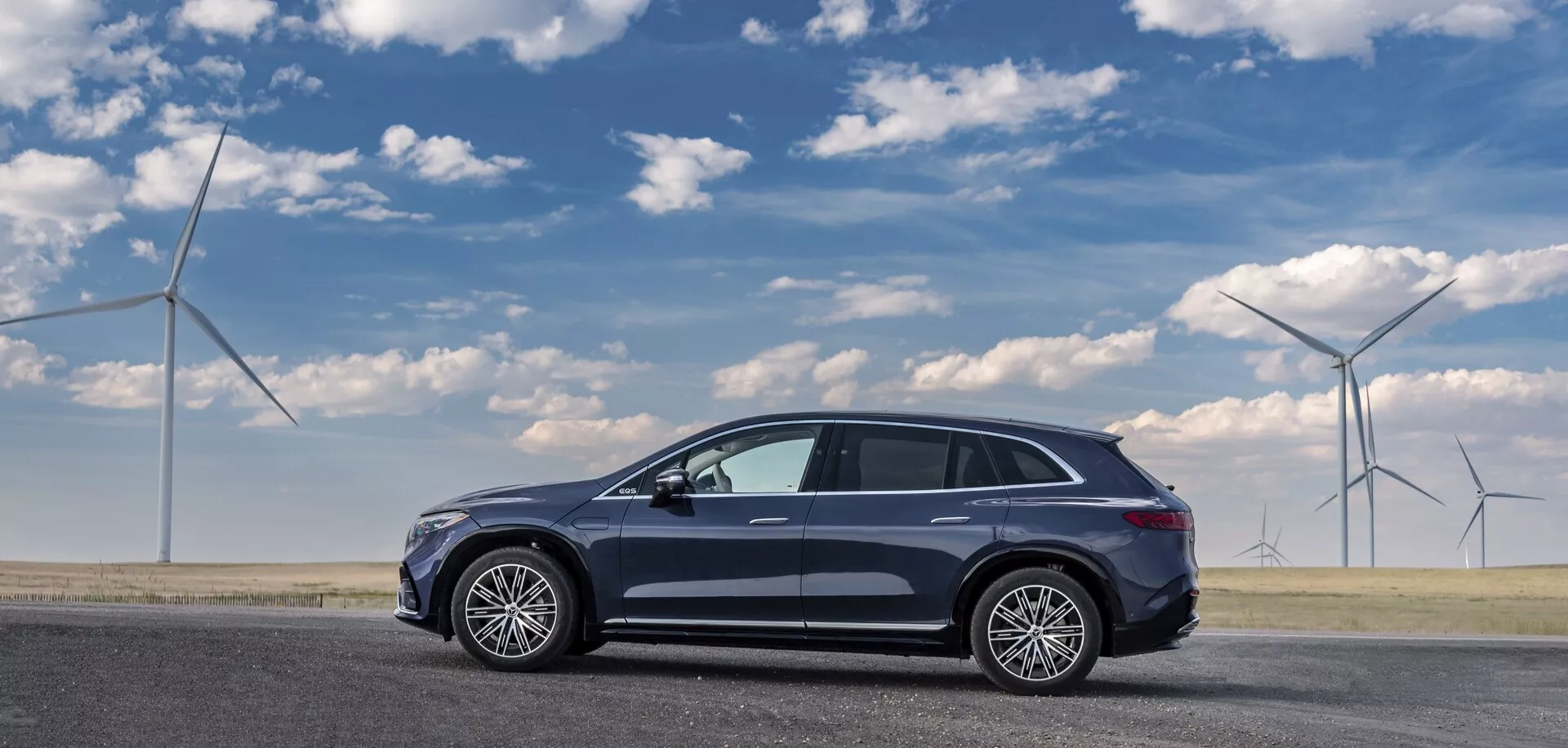Stranski pogled na temno obarvan Mercedes EQS-SUV 2022 parkiran na ravni cestni površini, ujet z voznikove strani. Sodobni crossover SUV aerodinamične in elegantne oblike ima pet vrat, velika platišča iz več krakov, panoramsko strešno okno, strešno anteno in bočna ogledala z vgrajenimi smerniki. Profil vozila nakazuje povišano držo, kar je značilno za crossoverje. Vetrne turbine v ozadju nakazujejo okolju prijazne teme.