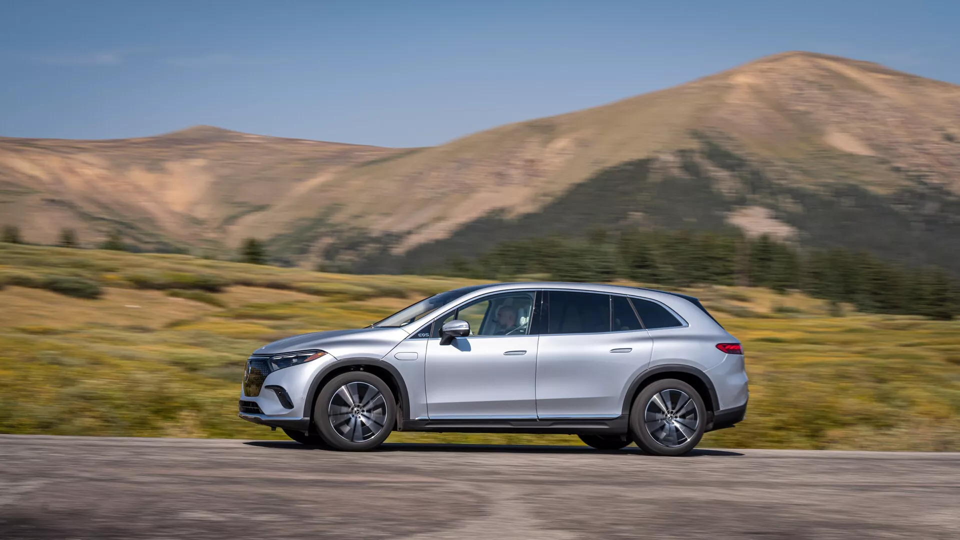 Mercedes EQS-SUV 2022 ujet v profilu, ki zavzema osrednji položaj v okvirju z gibom, vidnim iz rahlo zamegljenega ozadja. Velik SUV ima eleganten, zaobljen dizajn, aerodinamična bočna ogledala, lita platišča, strešne letve, kromirane obrobe in kovinsko barvo. Vidni so napredni tehnološki elementi, vključno z LED dnevnimi lučmi in poravnani ročaji vrat, kar nakazuje na brezključni dostop. Gorsko ozadje poudarja njegov dinamični in avanturistični videz, idealen za avtomobilske oglase in kontekste življenjskega sloga.