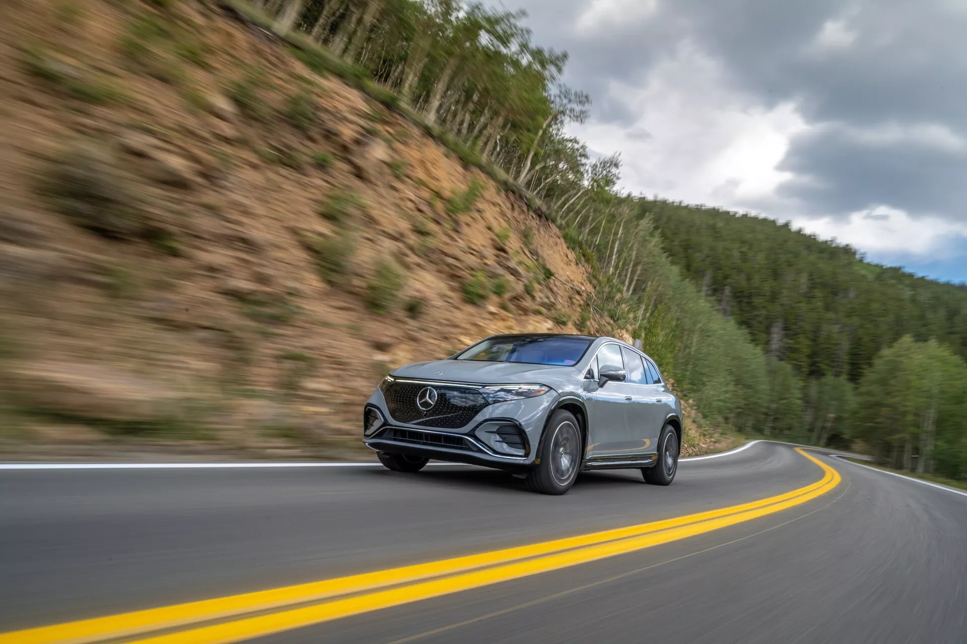Dinamičen posnetek Mercedes EQS-SUV 2022 v gibanju na gorski cesti, posnet iz nizkega, rahlo sprednjega kota. Avto, postavljen levo od sredine, se premika po zaviti dvopasovni cesti z zamegljenim ozadjem, kar kaže na visoko hitrost. Z elegantnimi LED žarometi, značilno sprednjo masko in kovinsko barvo se vozilo poudarja na ozadju gozdnatih gora pod oblačnim nebom. Podrobnosti, kot so zamegljena kolesa v gibanju in jasna bočna ogledala z morebitnimi vgrajenimi smerniki, povečujejo tehnološko privlačnost.